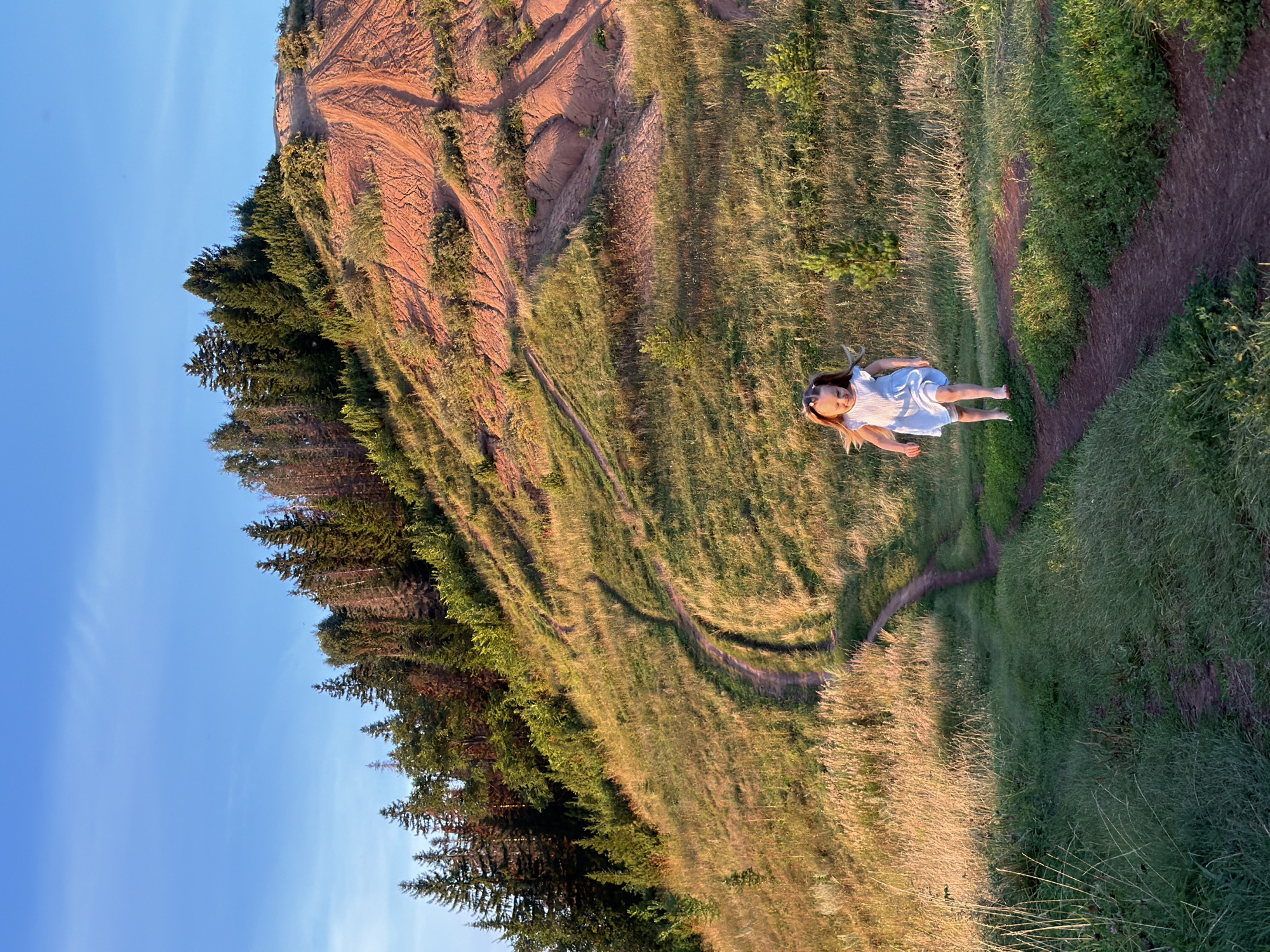 A young girl with blonde pigtails, wearing a white dress, runs barefoot on a dirt path winding through tall grass and low bushes. Behind her, the path ascends a steep, grassy hill dotted with young evergreen trees and leads to a reddish-brown, eroded hillside covered in sparse vegetation. The top of the hill is crowned with a dense forest of mature pine trees under a clear blue sky. The lighting suggests it is late afternoon or early evening, casting long shadows. The image appears to be taken in a natural, undeveloped area, likely a hillside or park on the outskirts of Olkhovka, Russia, given the presence of pine forests.