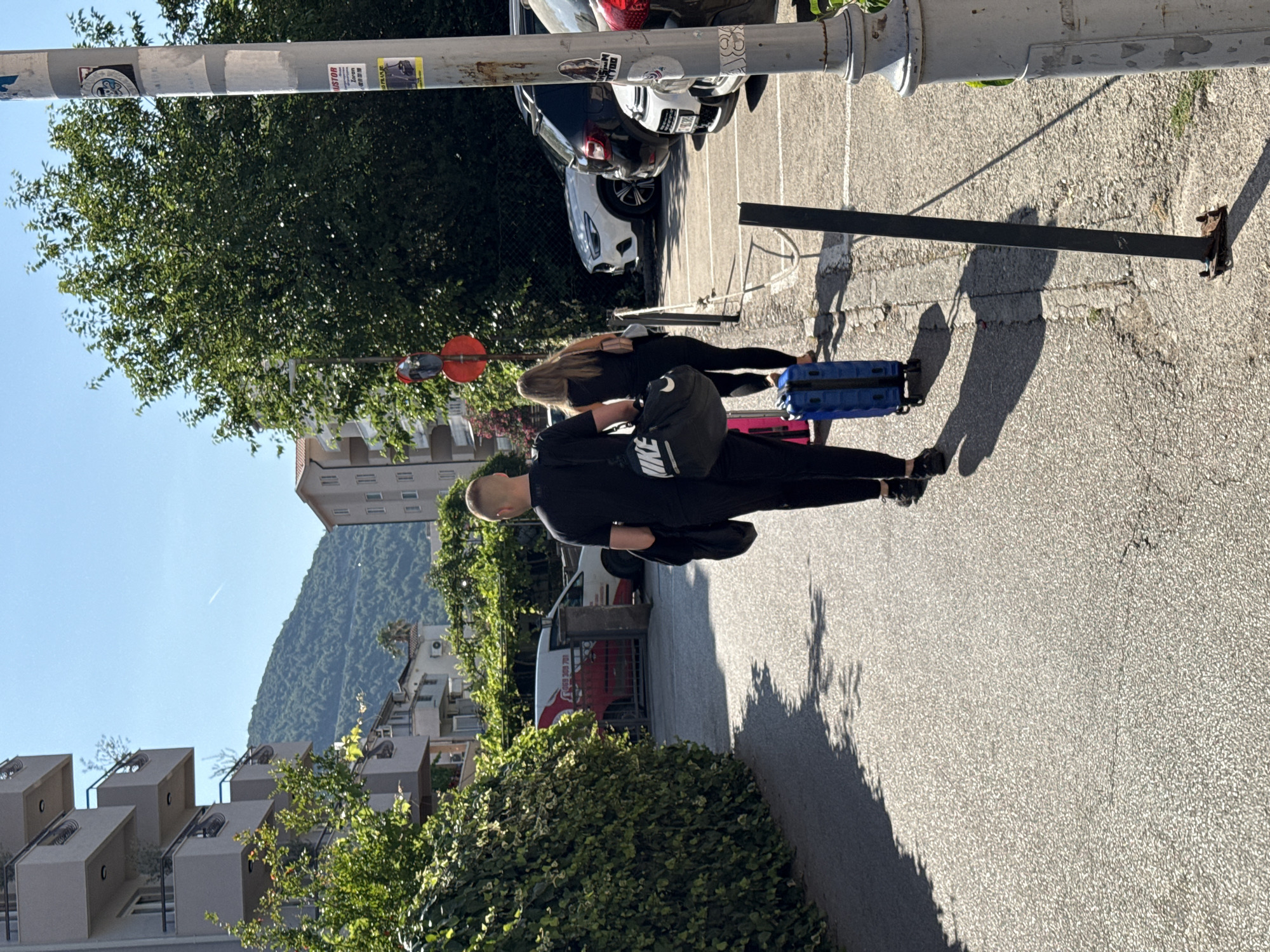 Two individuals, a man and a woman, walk away from the camera down a paved street in Budva, Montenegro. The man, on the left, wears a black t-shirt and black pants and carries a black duffel bag. The woman, on the right, wears a black top and black leggings and carries a light-colored purse. She is followed by a blue rolling suitcase. They are walking towards a parking lot where several cars are parked, including a white SUV and a black sedan. Buildings with balconies are visible on the left side of the street, and lush green trees line the area. In the background, a mountain covered in vegetation is visible under a clear blue sky. The scene appears to be a sunny day, with long shadows cast by the trees and the individuals.