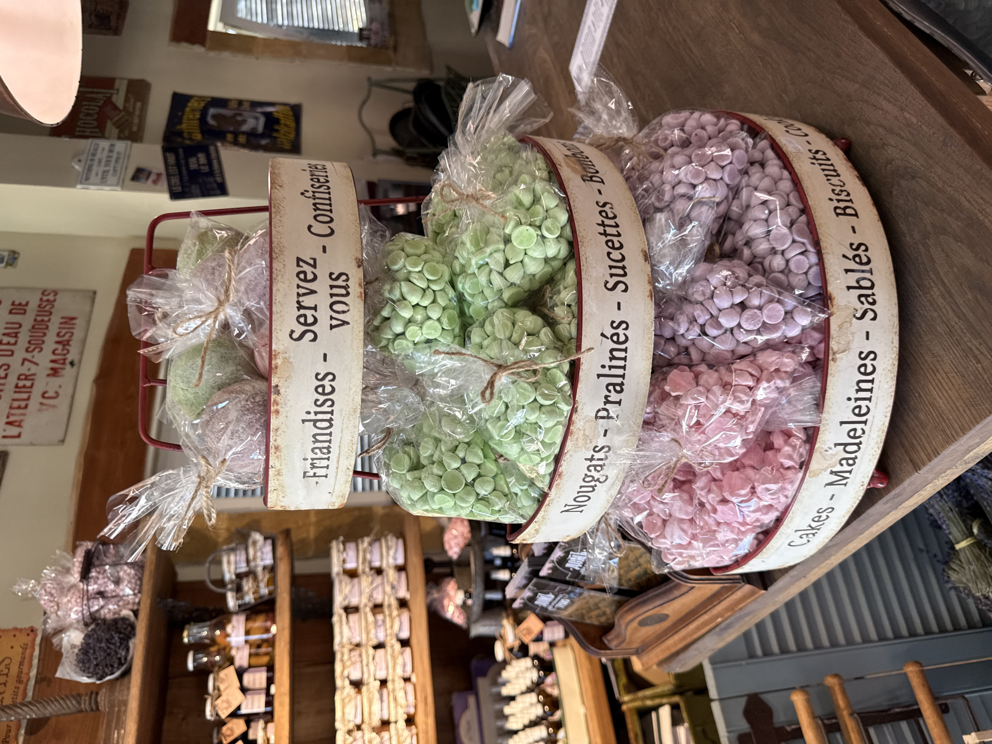 The image shows a tiered display of confectionery items in clear plastic bags, housed in red metal baskets with decorative rims. The rims are labeled with French text, indicating the types of sweets. The top tier contains purple and green sweets, labeled "Friandises - Servez vous - Confiseries." The middle tier has green sweets, labeled "Nougats - Pralinés - Sucettes - Bonbons." The bottom tier holds pink and purple sweets, labeled "Cakes - Madeleines - Sablés - Biscuits - Cookies."
The display is situated on a wooden counter in what appears to be a shop. In the background, shelves are visible, stocked with jars, bottles, and other packaged goods. Several signs with French text are also mounted on the wall behind the display. The overall setting suggests a French-style patisserie or confiserie. The image is well-lit, with the focus on the confectionery display. There are no people visible in the image.