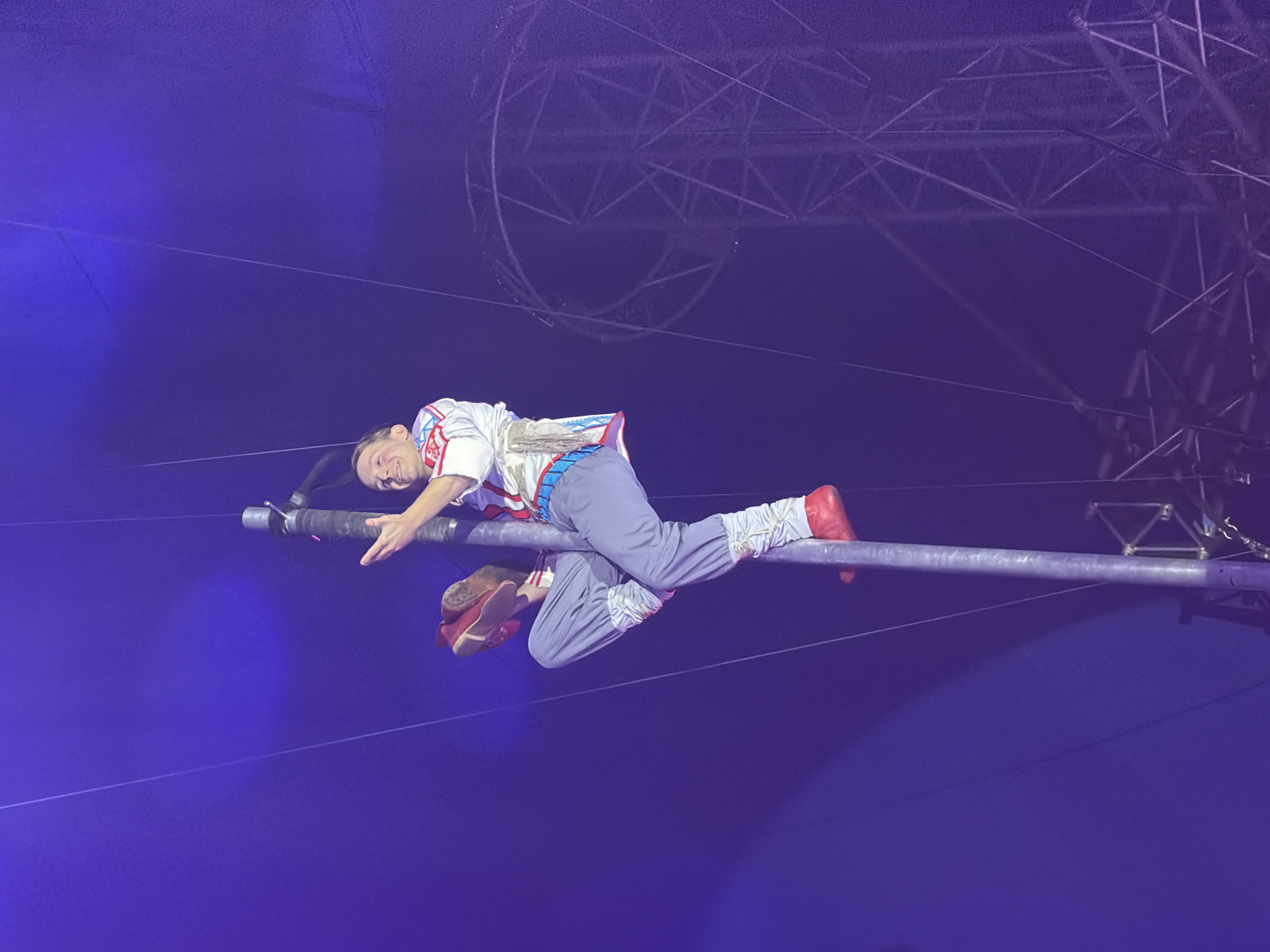 A male performer in a circus costume is lying horizontally on a trapeze bar, suspended in the air. He is wearing a white shirt with red and blue embellishments, gray pants, and red shoes. His left leg is bent, and his right leg is extended. His face is partially visible, and he appears to be smiling. The background is a dark, blueish-purple color, suggesting an indoor venue with dramatic lighting. Above the performer, there is a complex metal rigging structure, typical of a circus tent or arena. The overall impression is one of aerial acrobatics or a circus performance, highlighting the performer's balance and skill. The lighting is theatrical, with a focus on the performer and the trapeze bar.