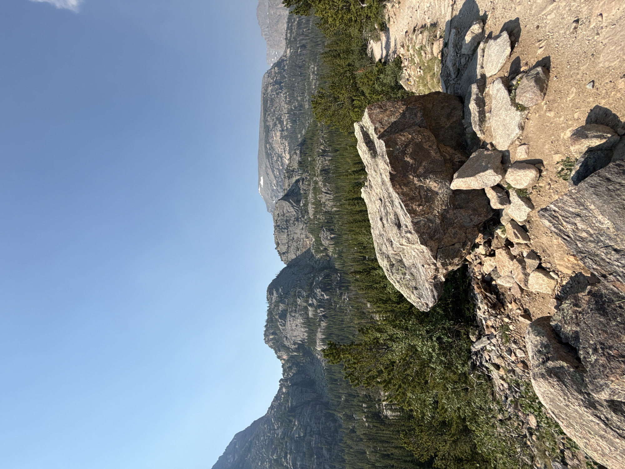 The image depicts a natural landscape with mountains, trees, and rocks. In the foreground, there are several large rocks scattered on a dusty, rocky path. A prominent boulder in the center foreground is a reddish-brown color and appears to be weathered. The path winds its way to the right and disappears into the distance. Behind the rocks and path, a dense forest of evergreen trees covers the lower slopes of the mountains. The mountains themselves are rocky and rugged, with some patches of lighter colored rock and vegetation visible on their surfaces. The sky above is clear and pale blue. The overall impression is of a remote and wild mountain environment. The location is likely within Rocky Mountain National Park, given the characteristic terrain and vegetation, and could be near Allenspark, Colorado, United States, a town bordering the park. There are no people or animals visible in the image, and no specific actions or interactions are depicted.