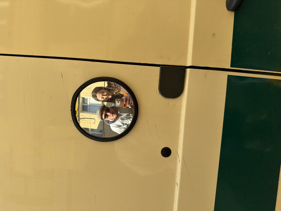The image captures a close-up of a small, round, black-framed convex mirror affixed to the beige-yellow painted surface of a vehicle. The vehicle's surface shows minor scuff marks and scratches, with a horizontal dark green stripe visible on its lower section. A black circular indentation and a black rounded rectangular stopper are present on the beige-yellow panel.
Reflected in the mirror are two individuals and a building facade. A male subject, positioned on the left, wears a grey flat cap, dark sunglasses, and a light-colored hooded jacket. He holds a light-colored smartphone with a prominent camera array extended in his right hand, appearing to capture a selfie. His mouth is slightly open. To his right and slightly behind him, a female subject with dark hair is visible, wearing a dark outer garment over an orange-brown top, also looking towards the smartphone.
The background reflected in the mirror reveals a multi-story building facade, rendered in a light yellow color, featuring multiple rectangular windows with white frames. This architectural style is characteristic of urban environments in Moscow, Russia, where the scene is situated.