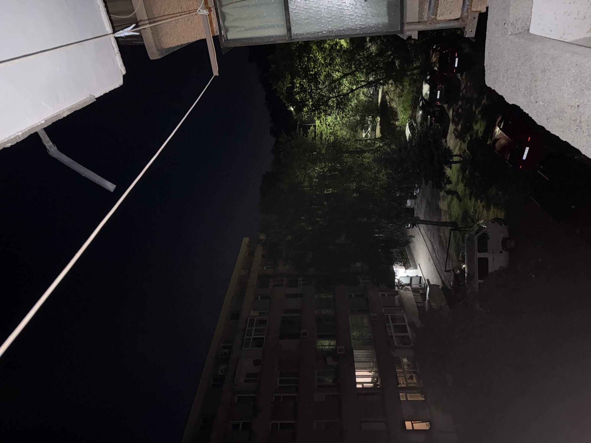This nighttime image, captured from an elevated perspective, depicts a residential area within the city of Kardzhali, Bulgaria, as indicated by the "KA" prefix on a visible license plate (KA 2198 BS).
In the immediate foreground on the right, a concrete balcony railing is partially visible. Extending horizontally across the upper frame is a clothesline, featuring a white plastic clothespin attached near the right side. Above the clothesline, an unfinished concrete balcony edge protrudes, and a bent, light-colored drain pipe extends downwards from beneath it. To the far right, a window with textured or frosted glass is visible, framed by a dark structure.
The central and left portions of the image feature a multi-story apartment building. The upper section of the building exhibits a light yellow facade, transitioning to a darker, possibly concrete or gray finish on its lower floors. Multiple windows are visible, with interior lights illuminated in some, notably a pinkish light on a lower-left window and a green light on a higher floor. Several external air conditioning units are mounted on the building's facade.
Between the apartment building and the background, a paved or unpaved area functions as a parking lot, bordered by numerous trees with dense foliage. A bright street lamp is visible in the middle distance among the trees, casting a strong, yellowish-green illumination on the surrounding leaves.
Several vehicles are parked in the area. A white van, consistent with a Volkswagen Transporter T3, is parked closer to the apartment building, equipped with a roof rack. Further into the parking area, multiple dark-colored passenger cars are visible. A red car in the lower-center features the Bulgarian license plate "KA 2198 BS". Other cars display plates from different Bulgarian regions, including "C 0081 MX" and "CB 0514 XT" (Sofia).
The background consists of a dark night sky. A discernible silhouette of distant hills or mountains, covered in dense vegetation, is visible against the horizon. No individuals are present in the visible scene.