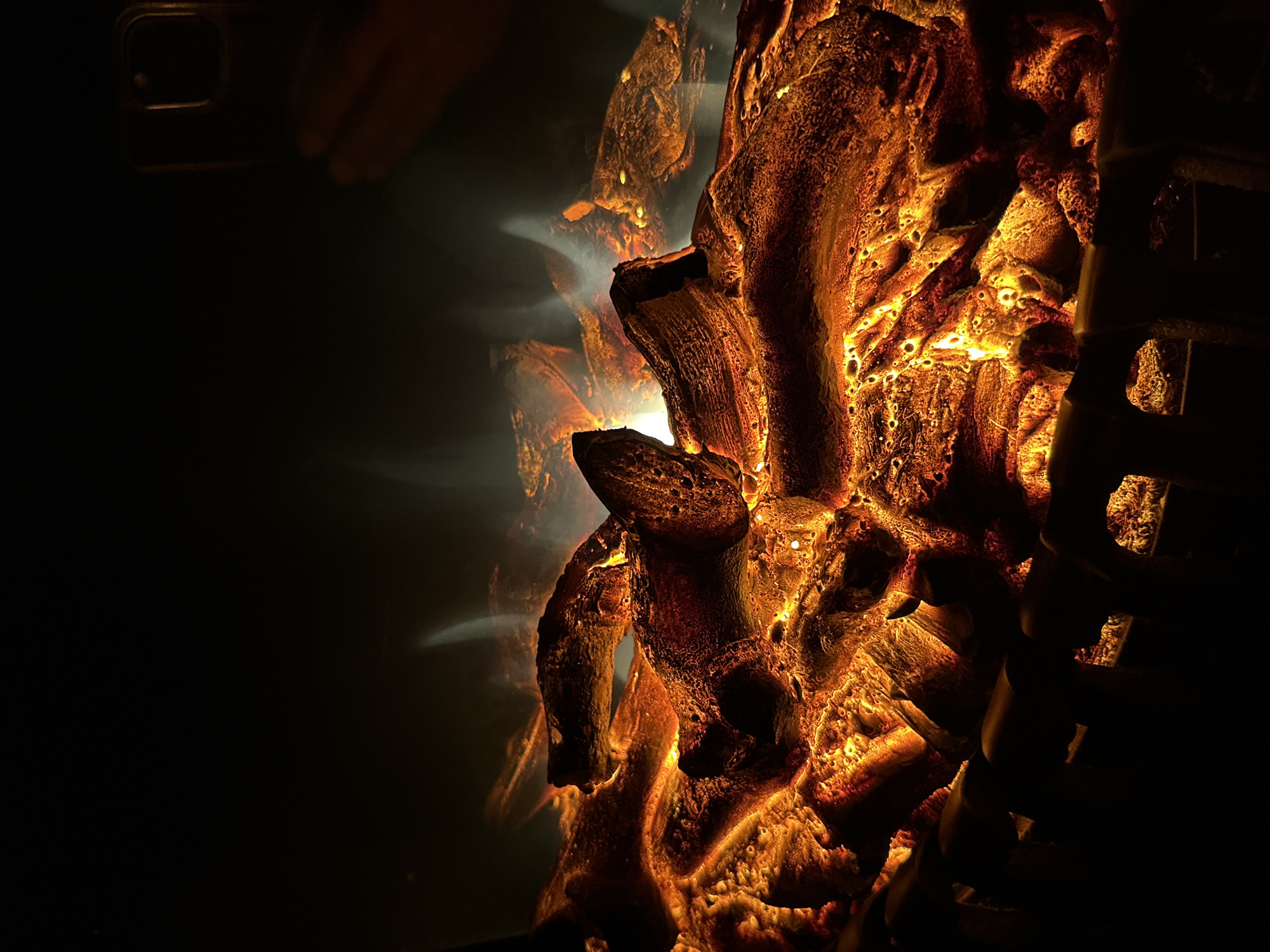 A close-up, dimly lit photograph captures the glowing embers and artificial flames of an electric fireplace. The scene is dominated by warm hues of orange, yellow, and red, emanating from the textured, log-like structures within the fireplace. Wisps of white and grey smoke-like material rise from the "embers," creating an illusion of active burning. In the upper right corner, a human hand is visible, holding a smartphone, suggesting someone is interacting with or documenting the scene. The overall environment appears to be indoors, with the background fading into darkness, emphasizing the fiery glow of the fireplace.