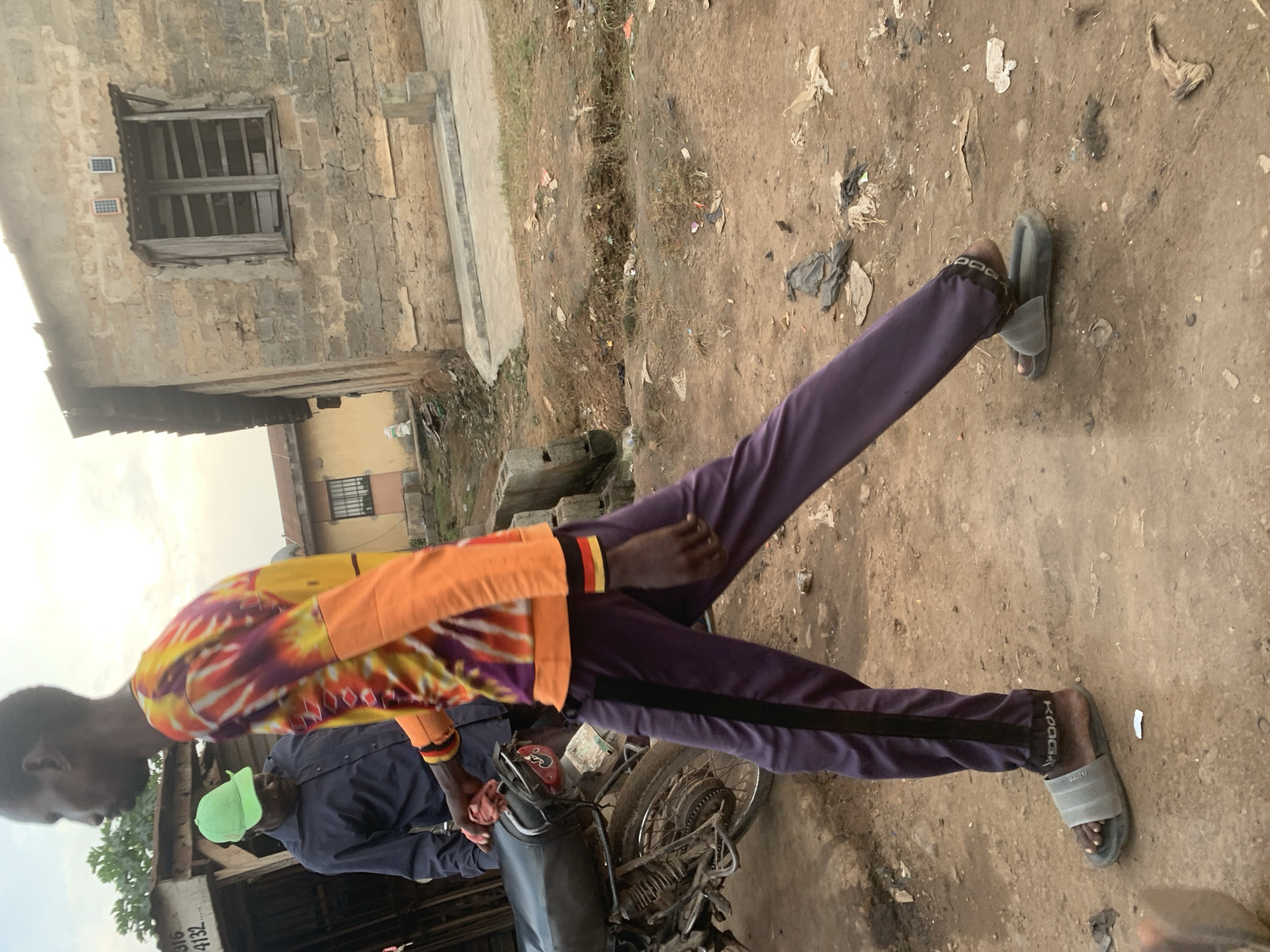 The image depicts a scene in Shagamu, Nigeria, characterized by a dirt road strewn with debris and unpaved surfaces. In the foreground, a person is walking, wearing a tie-dye long-sleeved shirt with orange, yellow, and purple colors, and dark purple pants with a black stripe. They are wearing grey slide sandals. To the left, a motorcycle is parked, and another person wearing a dark blue collared shirt and a bright green baseball cap is standing near it. Behind the people and the motorcycle are buildings constructed from concrete blocks. One building in the background features a corrugated metal roof and wooden window shutters, with two small solar panels mounted above the window. The overall environment appears to be a somewhat informal urban setting.
