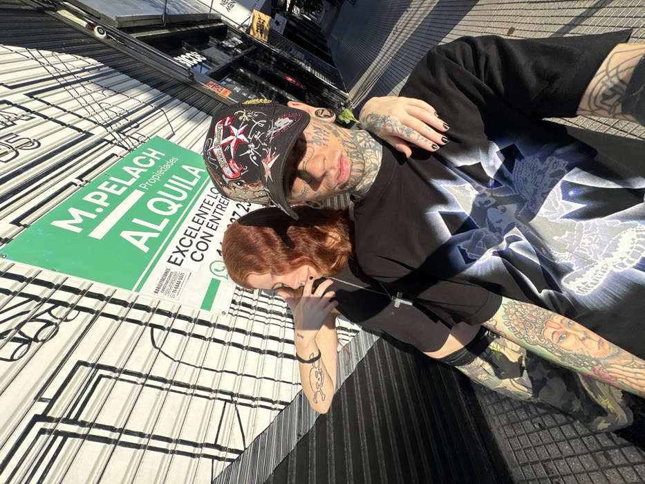 A person with tattoos and a cap is hugging a person with red hair. They are standing next to a green sign with white text that reads "M. PELACH Propiedades ALQUILA." The sign also includes contact information and the words "EXCELENTE LO CON ENTRER" and "27 23." They are standing in front of a building with a black and white striped awning. The people and the awning are slightly tilted in the frame.