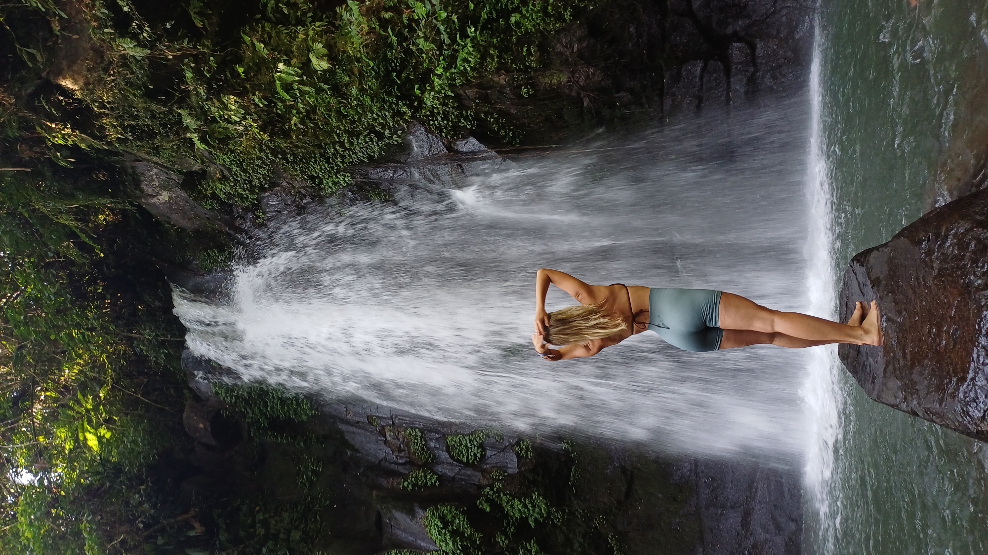 A medium shot, taken outdoors at what appears to be a waterfall in Tegallalang, Indonesia, shows a person, perceived as female, with her back to the camera. The subject, wearing a dark bikini top and blue shorts, is standing on a large, dark rock in front of the waterfall, facing it with her hands clasped behind her head, possibly stretching or posing. The waterfall cascades down from above, filling the frame with the motion and white spray of water. The surrounding environment is lush with green vegetation and dark rock formations, suggesting a tropical location. The image is rotated 90 degrees counter-clockwise.