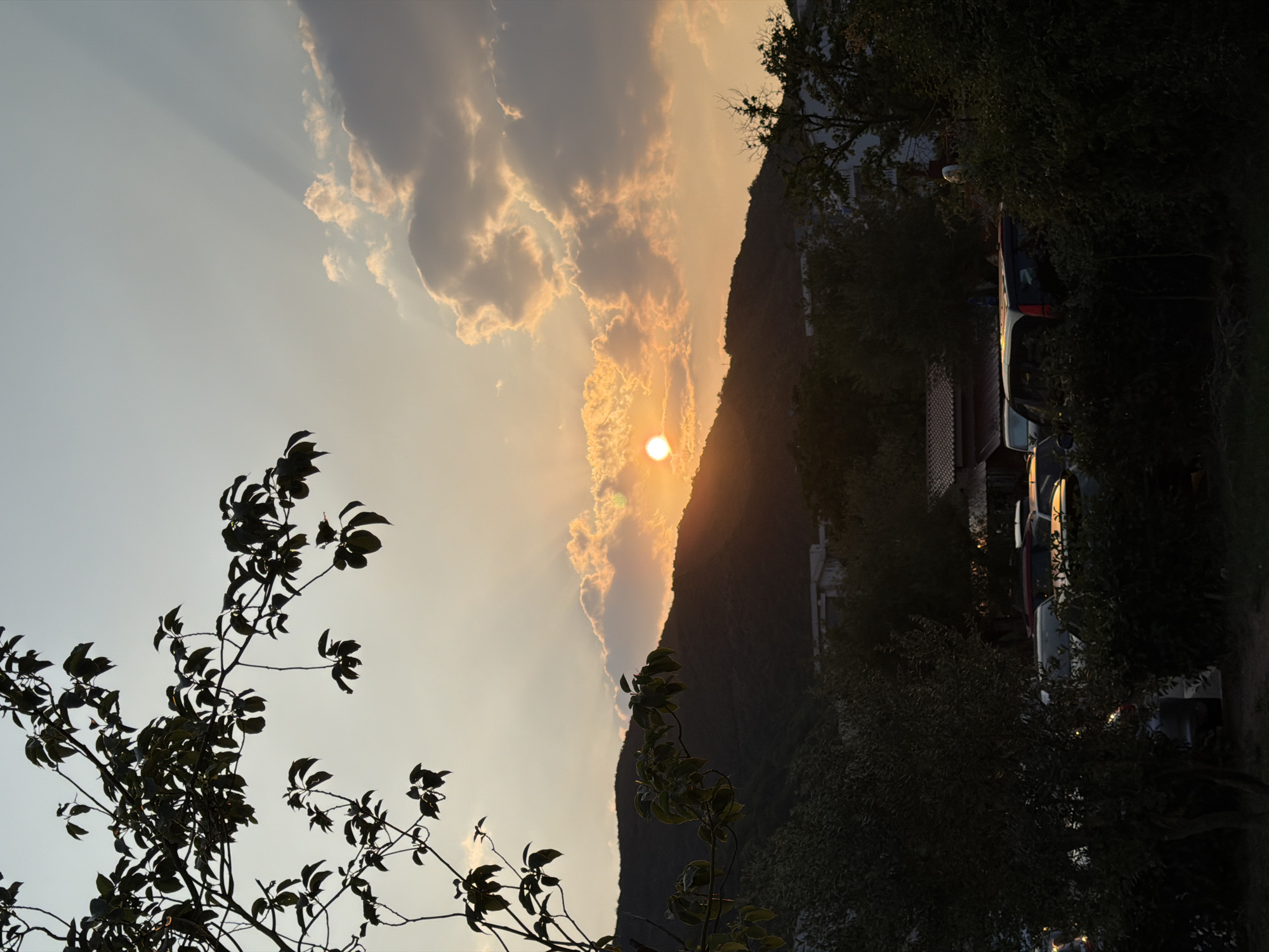 The image depicts a sunset over a hilly landscape, likely in the city of Budva, Montenegro. In the foreground, silhouetted trees with dark green leaves are prominent. Partially obscured by these trees, a collection of parked cars is visible. The cars are dark-colored and appear to be in a parking area.
The middle ground features a mountain range, silhouetted against the vibrant sunset sky. The sun itself is visible as a bright, orange orb, partially obscured by clouds and casting rays of light downwards. Some of the clouds are illuminated by the sunset, appearing in shades of orange and yellow. Buildings, possibly residential or commercial, are visible nestled at the base of the mountains.
The sky transitions from a lighter, pale yellow near the horizon to a soft blue at the top of the frame. The overall atmosphere is serene and captures the natural beauty of a sunset. There are no people or animals clearly visible in the image. The scene appears to be an outdoor setting, likely a residential or semi-urban area given the presence of cars and buildings.
