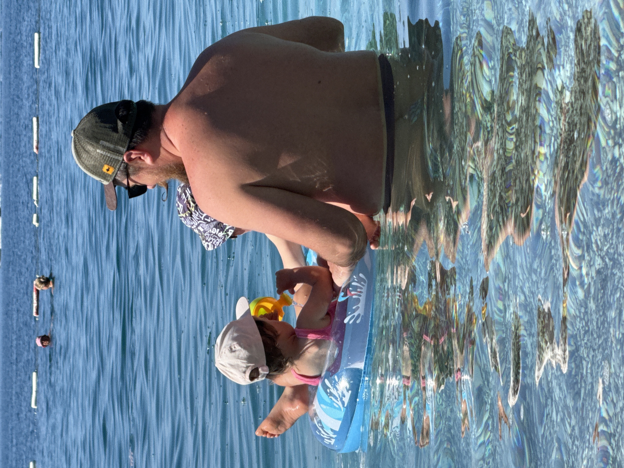 The image captures a scene in the clear, blue waters of Boreti, Montenegro, featuring an adult male interacting with two young children. The adult male, viewed from the back, is partially submerged in the water, wearing a dark green and brown mesh trucker hat with a small orange fish emblem on the left side, and dark sunglasses. He has a reddish-brown beard and his bare back and shoulders are visible. A young child, likely an infant or toddler, is seated within a blue inflatable swim ring with wave patterns, wearing a pink swimsuit and a light beige baseball cap. This child holds a yellow and orange plastic toy. A second small child, wearing a dark patterned hat, is situated between the adult male and the child in the float, with only their head and upper back visible. In the distant background, two other adults are visible in the water, beyond a line of several white rectangular buoys. The water is calm and highly transparent, revealing a light-colored, textured seabed beneath the surface. Light reflections and refractions are prominent on the water's surface.