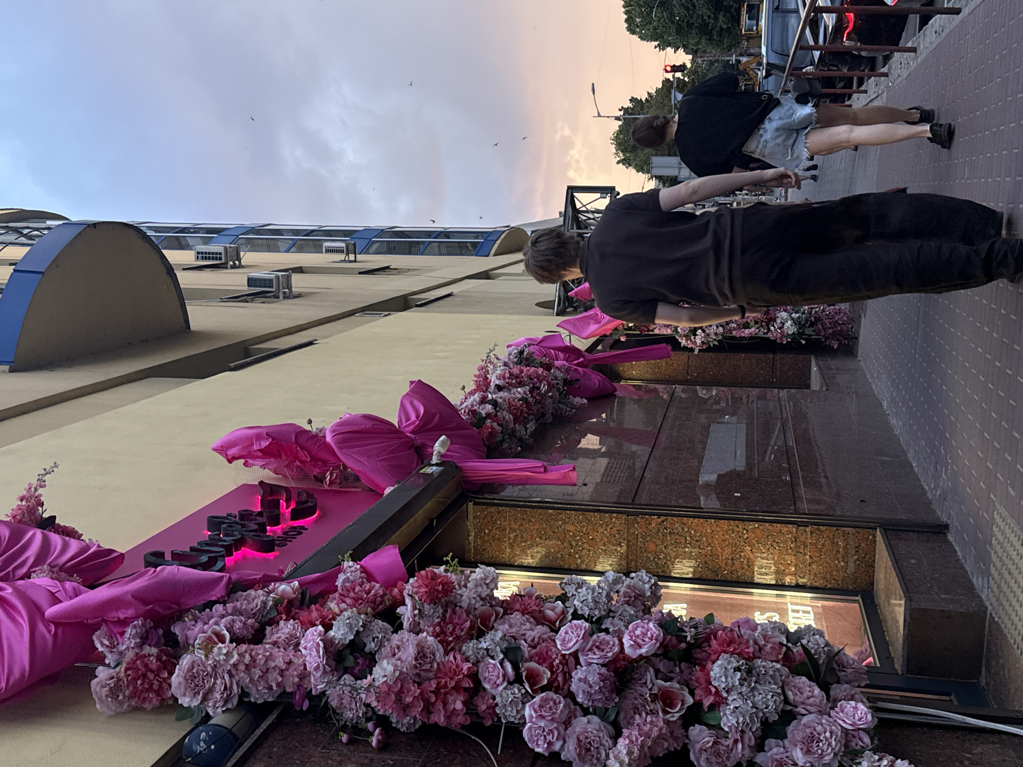 This is a medium shot, eye-level, outdoor, daylight, street-level photograph taken on a cloudy day in the city of Dnipro, Ukraine. The scene depicts a storefront decorated with abundant pink artificial flowers and draped pink fabric. To the right of the storefront, two individuals, a man and a woman, are walking away from the camera on a paved sidewalk. The man is wearing a dark t-shirt and dark, loose-fitting pants. The woman is wearing a black t-shirt and distressed denim shorts. Behind them, a multi-story building with a rounded blue balcony is visible. The sky is a mix of pink and grey clouds, indicating either dawn or dusk. A traffic light is visible in the distance. The overall atmosphere is one of urban commercial activity.