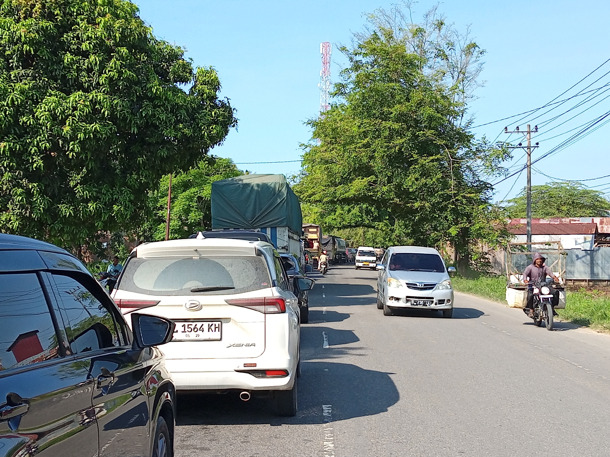 In Blangpanjoe, Indonesia, a traffic jam unfolds on a paved road under a clear blue sky. The foreground is dominated by the side of a black car, reflecting the surrounding environment. Behind it, a white SUV, identified by the license plate "L 1564 KH," is stationary. Further down the road, a silver van is also stopped, and beyond that, a line of trucks, one with a green tarp covering its load. A lone motorcyclist, wearing a hooded jacket, navigates the traffic, carrying bags on the back of his motorcycle. Tall green trees line both sides of the road, and in the distance, a tall communications tower is visible. Utility poles with numerous wires are also present. The scene depicts a typical day with moderate traffic on a road in Blangpanjoe.
