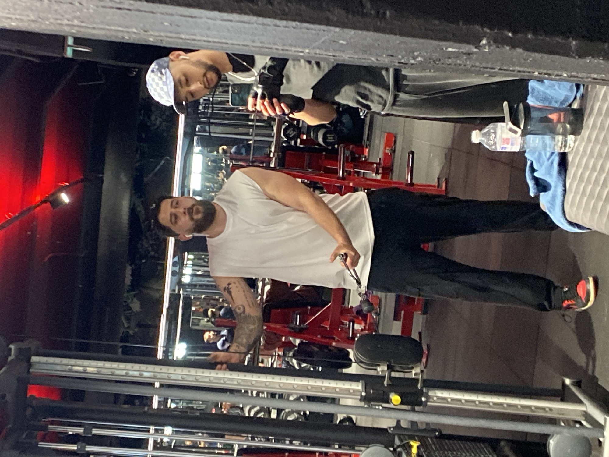 The image shows two men inside a gym in Strasbourg, France. The man in the foreground, wearing a white t-shirt and black pants, is using a cable machine. His left arm is sleeved with tattoos. The second man, wearing a gray sweatshirt and a white-and-blue patterned cap, is observing and holding a smartphone. There is a water bottle and a blue towel on a seat nearby. The gym is equipped with red-colored weightlifting machines.