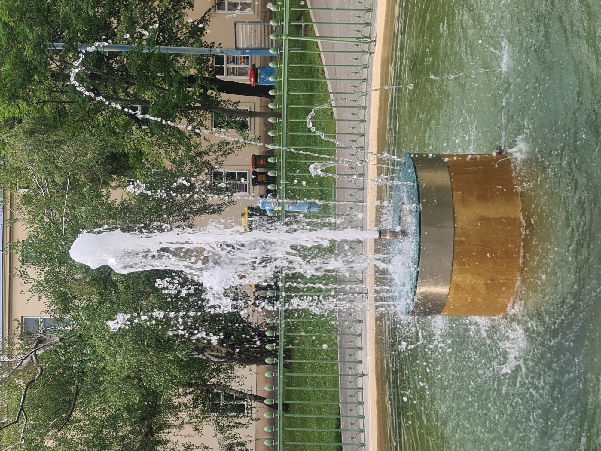 A photograph captures a fountain in an urban setting. The fountain features a cylindrical base with a gold-colored exterior and a silver metallic band at the top, from which water shoots upwards in multiple streams, some forming arcs. The water sprays create a dynamic visual effect, with droplets and splashes visible. The fountain is situated in a shallow pool with murky green water.
Behind the fountain, a green metal fence with decorative elements runs horizontally, separating the fountain area from a grassy patch and a paved walkway. In the background, several trees with dense green foliage provide a natural backdrop. Beyond the trees, a light-colored building with multiple windows is visible, suggesting an urban environment. Some rubbish bins are also present near the building. The scene appears to be an outdoor public space. The image does not contain any discernible people. The location is identified as Vienna, Austria.