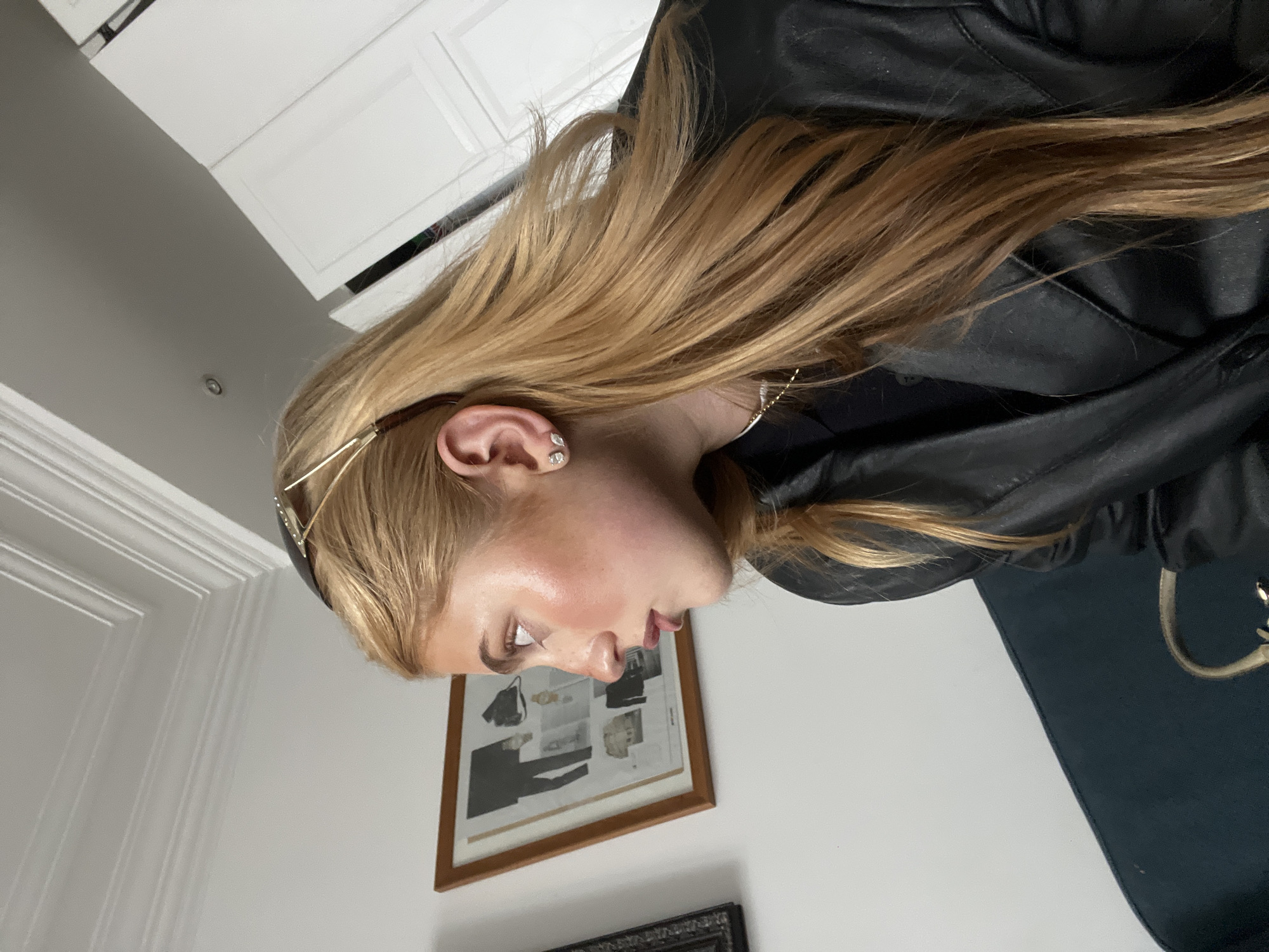 A woman with long, blonde hair, wearing a black leather jacket and a gold headband, is positioned against a wall in what appears to be an indoor space. The wall behind her is light-colored and features two framed images. The lower image is partially visible and has a dark frame, while the upper image, framed in brown, contains a collage of fashion sketches and photographs. A partially visible white cabinet is in the upper left corner of the frame, and a dark-colored piece of furniture is present in the lower right. The setting appears to be a residential or commercial interior rather than an outdoor location in Paris, France.