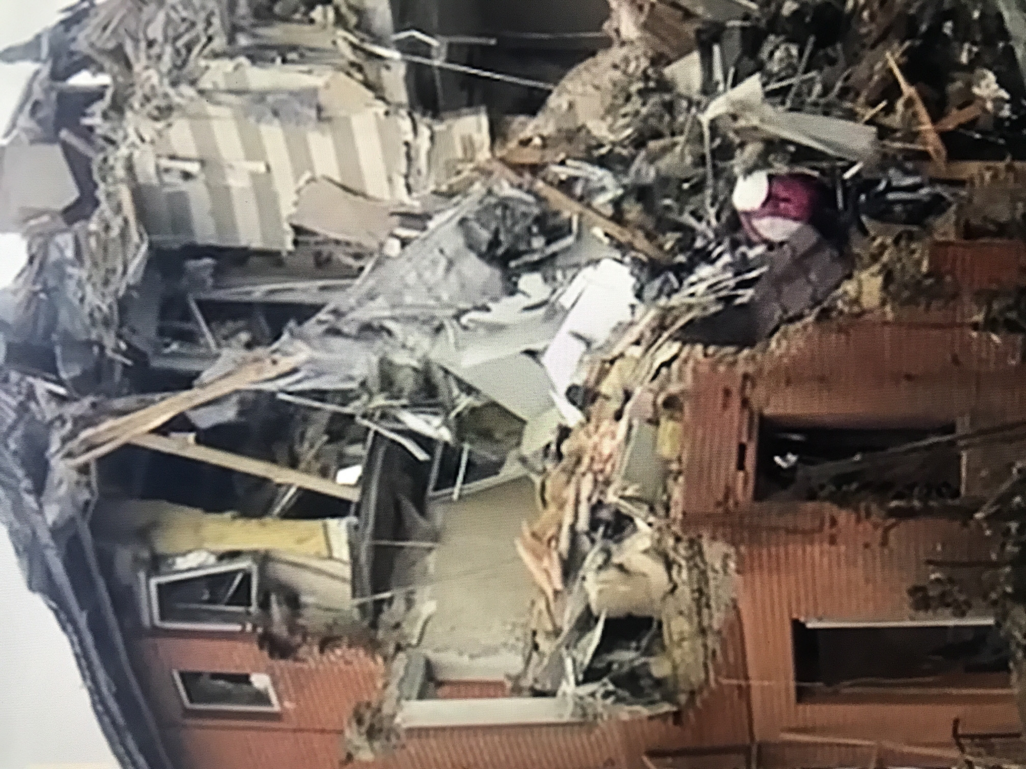 The image shows a collapsed building in Donji Pobori, Montenegro. The structure is heavily damaged with rubble and debris scattered around. There are visible sections of the building's facade with window frames. A pink and white object, possibly a toy or personal item, is visible amidst the debris. The overall scene suggests a disaster, potentially caused by an earthquake, explosion, or structural failure. There are no people visible in the image.