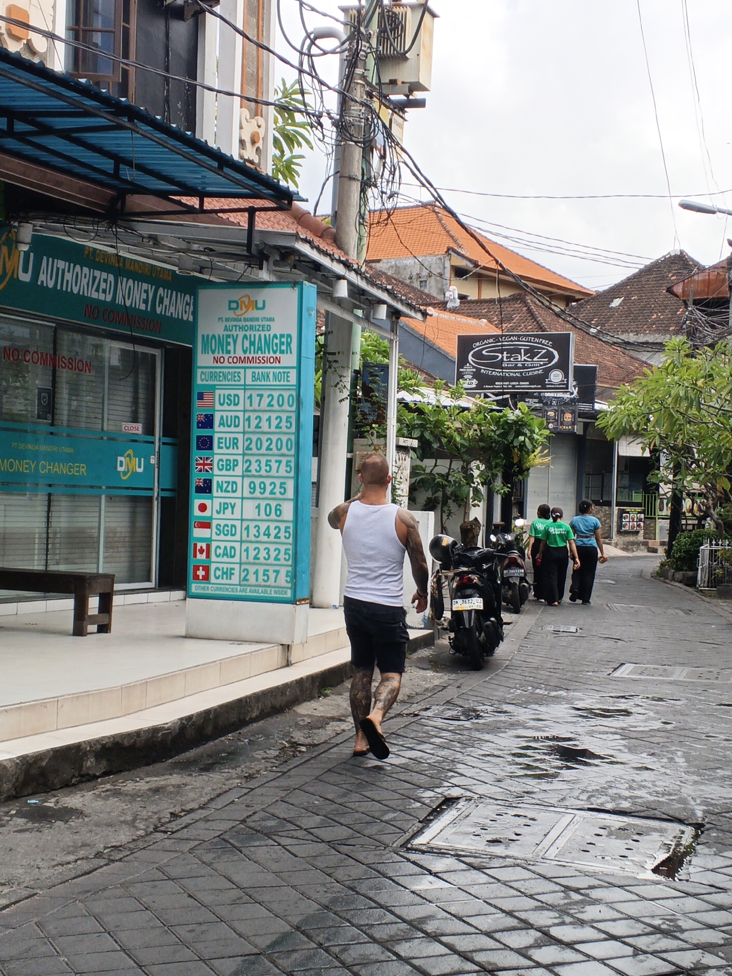 The image depicts a street scene in Kuta, Indonesia. On the left, a money changer establishment with a teal awning and signage reading "PT. DEVINA MANDIRI UTAMA AUTHORIZED MONEY CHANGE NO COMMISSION" is visible. A price board displays currency exchange rates, with USD at 17200 and EUR at 20200, among others. A wooden bench sits in front of the establishment. A man with tattoos on his legs, wearing a white tank top and black shorts, walks away from the camera down the paved street. To the right, two women in green shirts and one woman in a blue shirt walk away from the camera. Several motorcycles are parked on the side of the street. The street is wet with puddles, suggesting recent rain, and is paved with dark square tiles. Buildings with red tile roofs are visible in the background, along with overhead electrical wires. A sign for "StakZ Bar & Grill" is also visible. The sky is overcast.