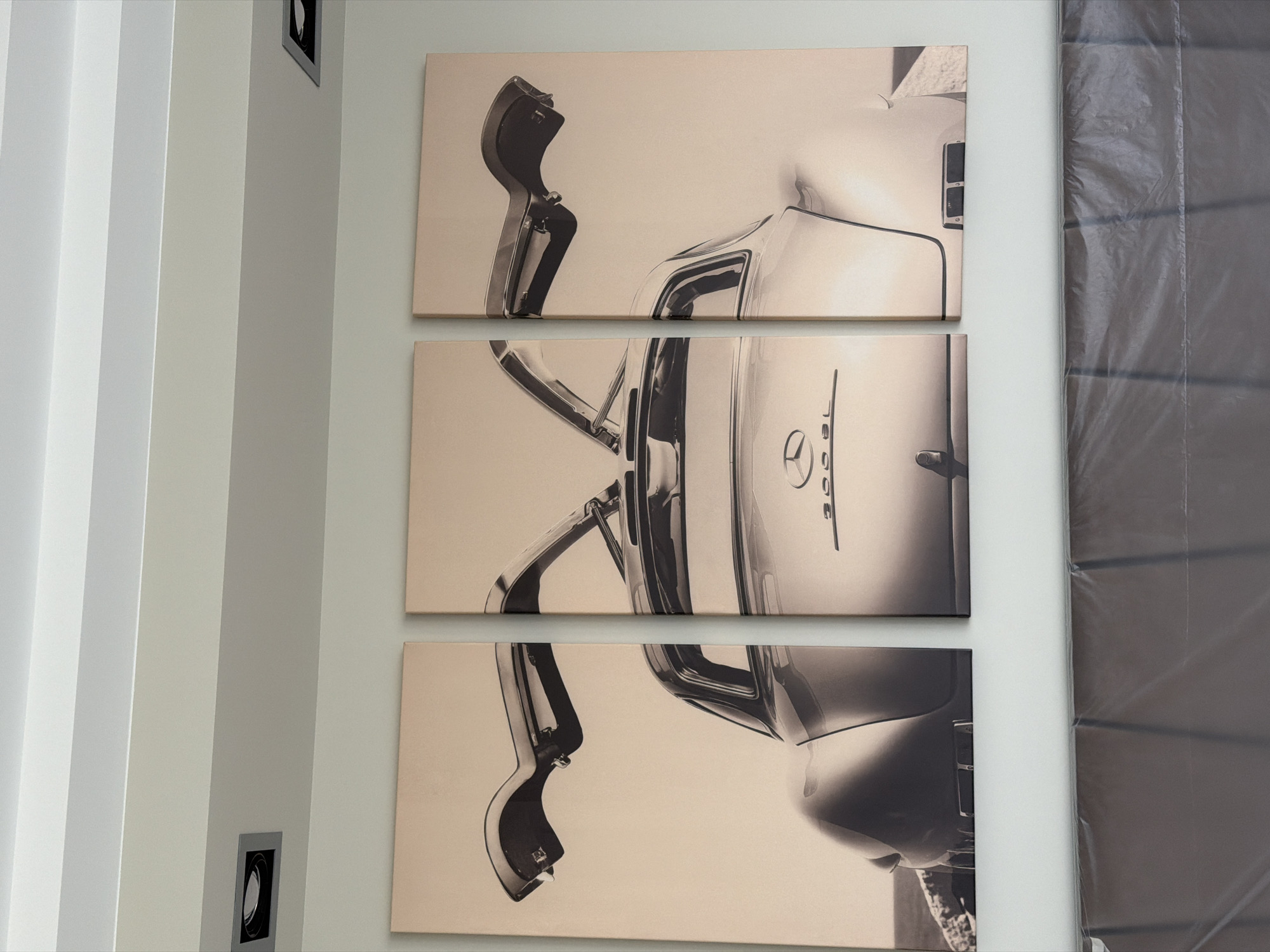 A triptych artwork depicting the rear view of a vintage Mercedes-Benz 300 SL Gullwing is mounted on a white wall in an indoor setting. The artwork is split into three vertical panels of equal size, with the car's rear end and open gullwing doors dominating the composition. The left panel shows the left gullwing door in its upward position and a portion of the car's rear. The middle panel displays the central rear of the car, including the Mercedes-Benz logo and the "300 SL" badging. The right panel shows the right gullwing door in its open position, with a hint of the car's rear quarter panel. The artwork has a sepia tone, giving it a vintage aesthetic. Two recessed square lights are visible on the white ceiling above the artwork. The bottom portion of the image shows a draped object, possibly furniture covered in protective plastic, in front of a gray wall. The location is identified as Khimki, Russia.