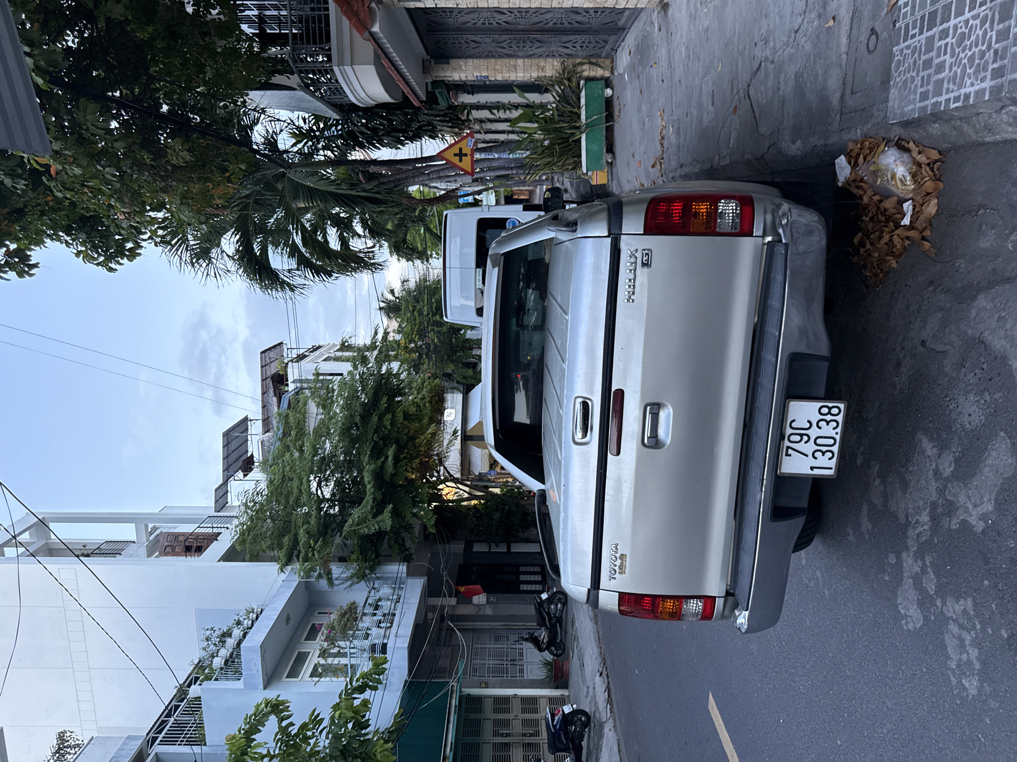 The image depicts a street scene in Nha Trang, Vietnam. In the foreground, a silver Toyota Hilux pickup truck is parked on the right side of the road, facing away from the camera. The truck's license plate reads "79C 130.38". To the right of the truck, a sidewalk is visible, with a green planter box containing plants and a yellow traffic sign with a cross symbol. Further down the street, a white van is parked. On the left side of the image, there are several buildings with balconies and foliage. A small Vietnamese flag is hanging from one of the buildings. The sky is overcast, and the lighting suggests it is late afternoon or early morning. The street appears to be a residential or mixed-use area, with a mix of modern and older architecture. A pile of dried leaves and trash is visible on the ground near the rear of the truck. Two motorcycles are parked on the sidewalk in front of a building on the left.