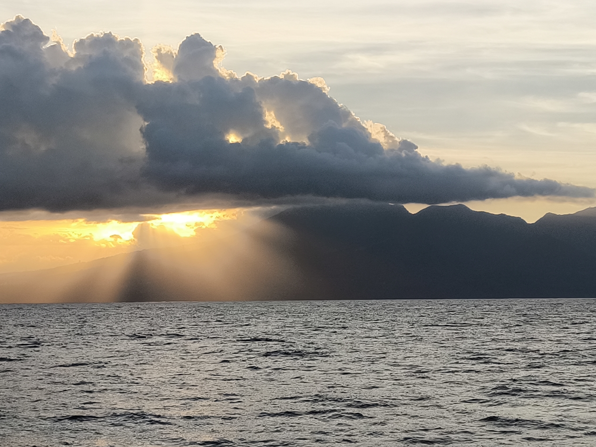 The image shows a seascape at sunset in an unknown city of Indonesia. A body of water with small waves covers the foreground. In the midground, there is land with low mountain-like terrain in the middle and to the left. A heavy, dark cloud formation sits above the land, obscuring parts of the land. The sun is positioned behind the land and the cloud, and sun rays radiate downward, piercing through breaks in the clouds. The sky is a mix of colors from white to grey to orange and yellow, reflecting the sunset. There are no people visible in the scene.