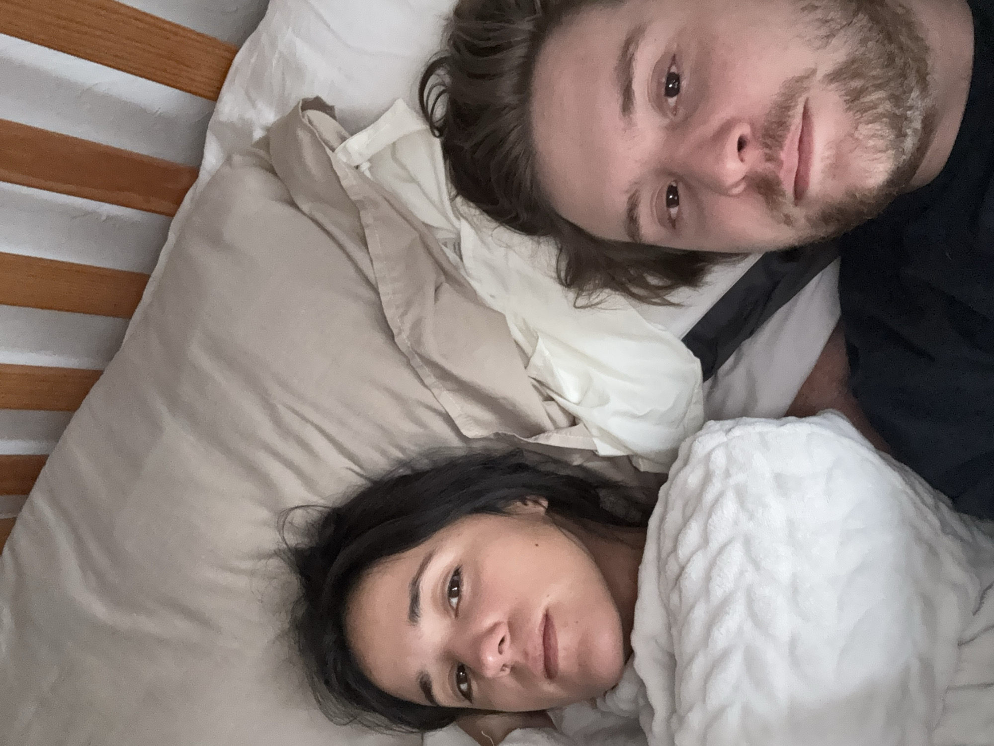 A selfie shot from an elevated perspective depicts two individuals lying in a bed. The male subject, positioned on the top right, has fair skin, dark brown hair, and a beard. The female subject, located on the bottom left, has olive skin and dark hair. Both appear to be looking directly at the camera. They are partially covered by bedding. The setting is a bedroom, featuring a wooden bed frame. The location cannot be identified as being in Mexico City, Mexico.
