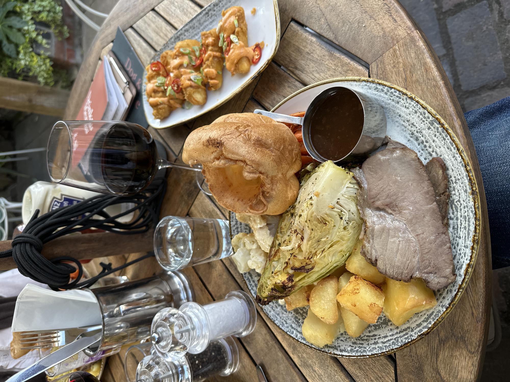 On a wooden table, a plate of roast dinner and a side of food are arranged. The roast dinner includes slices of roast meat, roasted potatoes, cabbage, cauliflower, a Yorkshire pudding, gravy in a small metal container, and possibly carrots. The side dish consists of fried food items topped with a sauce and chili slices. Nearby are a wine glass filled with red wine, a glass of water, salt and pepper grinders, cutlery, and what appears to be condiments and a black rope. The photo appears to be taken outdoors at a dining establishment in West Suffolk, United Kingdom.
