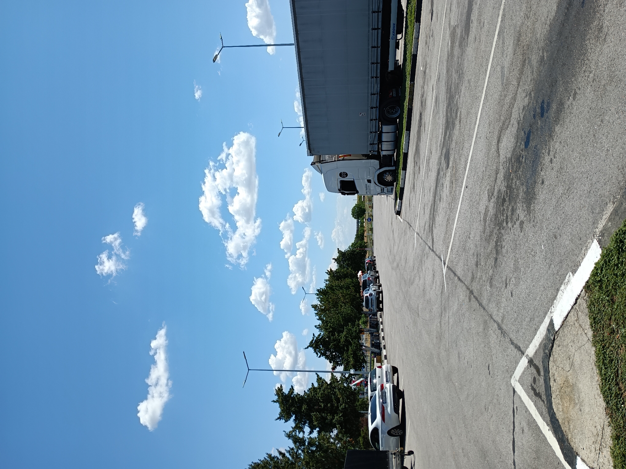 A bright, sunny day is depicted in Radinovo, Bulgaria, with a clear blue sky dotted with fluffy white clouds. The scene is a parking lot, likely at a truck stop or rest area, given the presence of a large semi-trailer truck parked on the right side of the frame. The truck has a white cab and a light gray trailer. In the background, several cars are parked in a row, and more trees are visible. The ground is paved asphalt, with white parking lines and some dark oil stains. Streetlights are visible in the distance, along with more trees and some low vegetation. The perspective suggests the photo was taken from ground level, looking towards the parked vehicles and the horizon.