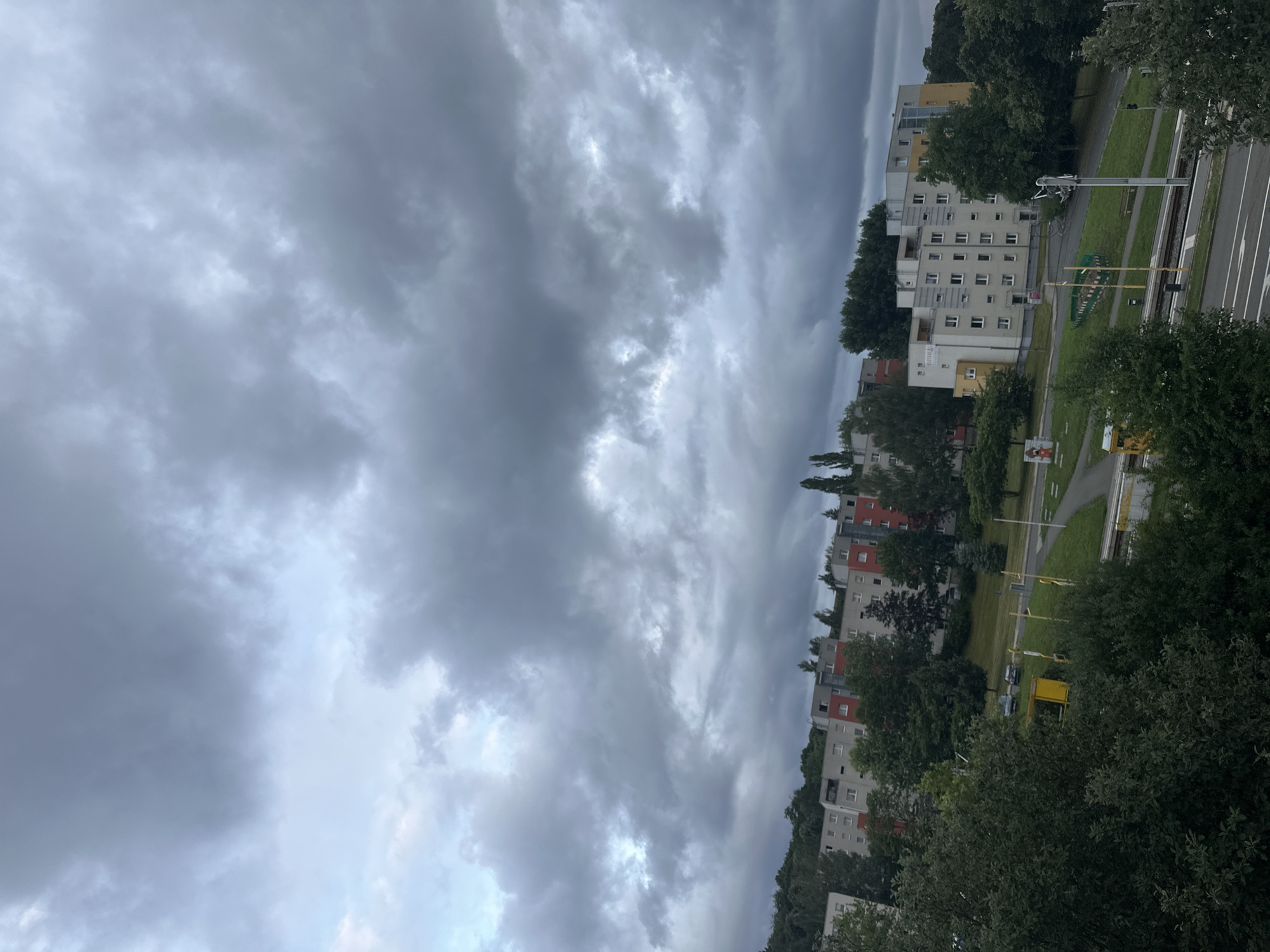The image depicts a cloudy sky over a residential area. In the foreground, dense green trees obscure the lower portion of the scene. Beyond the trees, a row of multi-story apartment buildings with varying facade colors, including white, grey, red, and yellow, are visible. The buildings appear to be part of a post-war housing development. Adjacent to the buildings is a grassy slope with scattered trees. A road with traffic markings runs through the scene, and a tram track is visible. Several streetlights and a yellow structure are also present. Two cars are parked on a grassy area between the buildings and the road. The overall atmosphere is overcast due to the thick, dark clouds.
