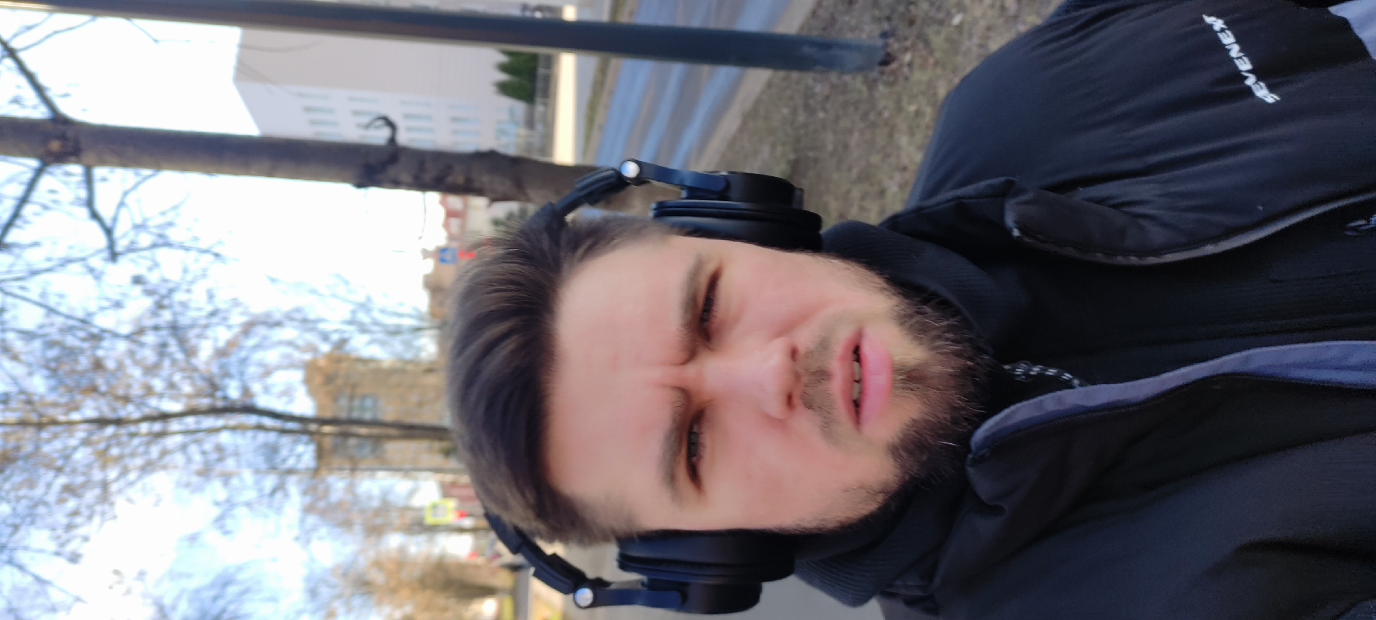 A selfie image shows a man wearing headphones and a black puffer jacket with the word "SEVENEXT" visible on the shoulder. The man has fair skin, dark hair, and a beard. The background shows an urban landscape with trees and buildings, suggesting a street scene in the city of Fryazino, Russia. The sky is overcast. A utility pole is visible to the left.