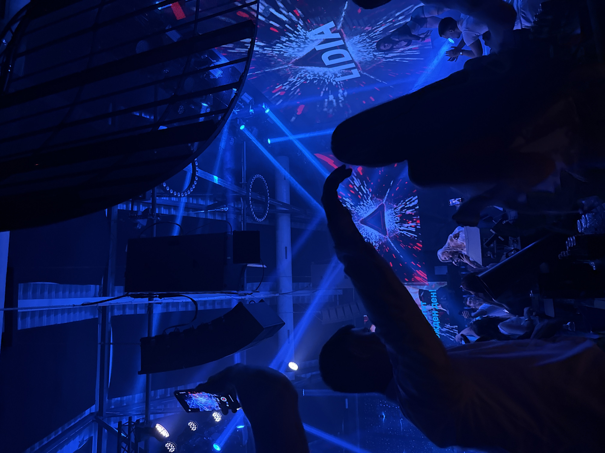 The image depicts a lively indoor event, likely a nightclub or concert venue, bathed in vibrant blue and red lighting. Silhouettes of attendees are visible, with some holding up their phones, suggesting they are capturing the moment. In the background, a large screen displays the word "LIDIA" in bold white letters amidst a dynamic, abstract visual. A performer, possibly a singer, is seen on a stage in the middle ground, illuminated by spotlights. The environment appears to be a dimly lit space with prominent stage lighting and sound equipment, creating an energetic and immersive atmosphere.