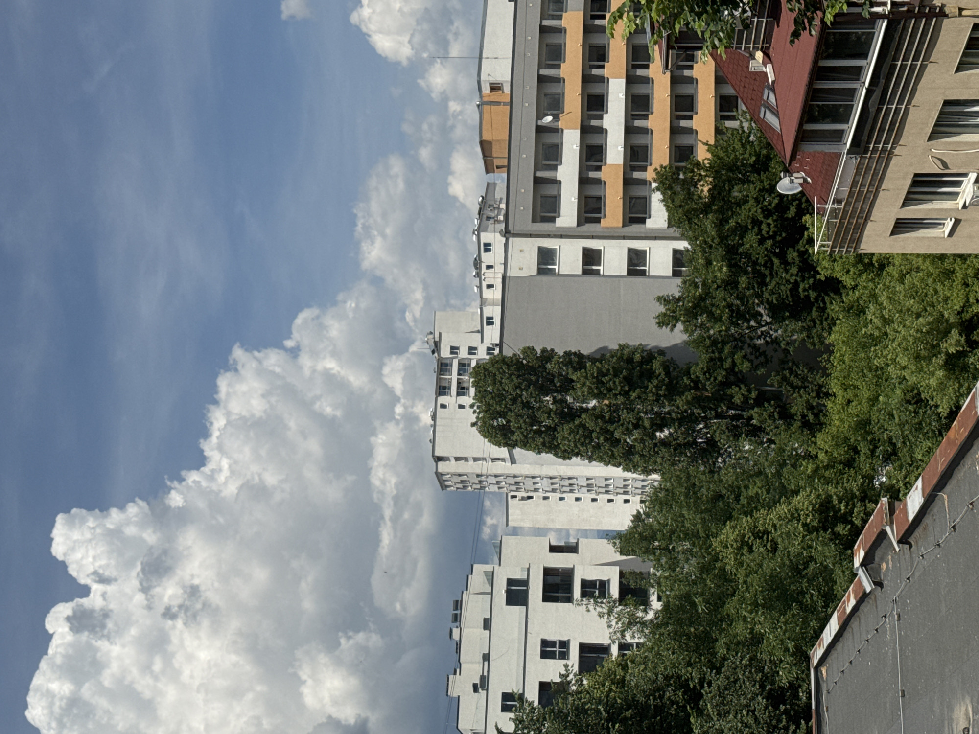 The image shows a cityscape with multiple buildings and lush green trees. The buildings are a mix of modern apartment blocks and older structures, with varying architectural styles and colors. Some buildings are tall and white, featuring multiple windows and balconies, while others are shorter with orange and grey facades. The foreground features the edge of a dark-colored rooftop with weathered railings. Dense green trees are prominent throughout the scene, partially obscuring some of the buildings. The sky is blue with large, fluffy white clouds. The scene appears to be taken from an elevated vantage point, looking across the urban landscape. Based on the architectural style of the buildings, particularly the apartment blocks with their repetitive window patterns and the presence of mature trees in a dense urban setting, this scene is consistent with areas in Sofia, Bulgaria, such as the Mladost or Druzhba districts, known for their large residential complexes. There are no people or animals visible in the image. The time of day appears to be daytime, with clear sunlight casting shadows. The overall atmosphere is one of a developed urban environment with significant greenery.