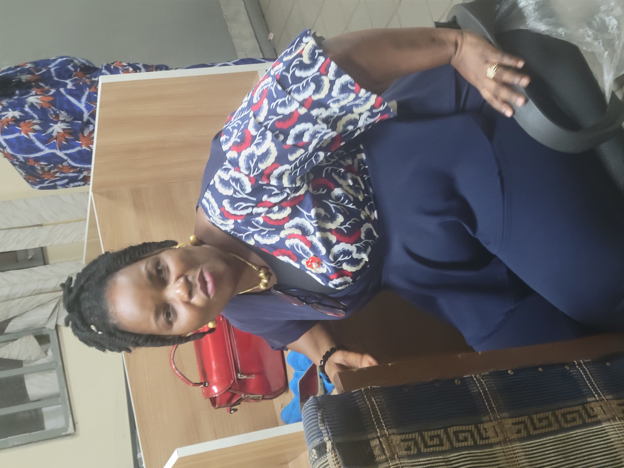 A woman is seated in an office chair within an interior space, possibly in Abuja, Nigeria. She is wearing a dark blue dress with a floral patterned collar. The office setting includes a wooden desk, a red handbag, and an upholstered object with dark blue and tan patterned fabric. A window with light filtering through is in the background.