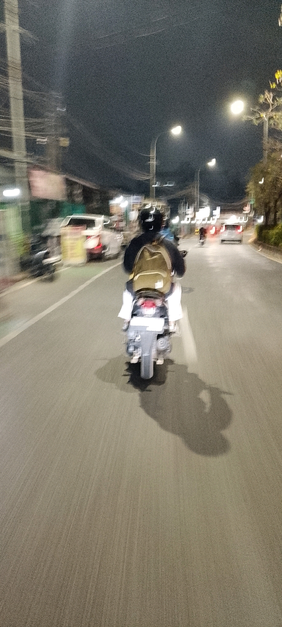 A person rides a motorcycle at night through the city of Bekasi, Indonesia. The rider, wearing a helmet and a backpack, is moving away from the camera. The scene is characterized by motion blur, indicating the motorcycle is in motion. Streetlights illuminate the road and surrounding environment, casting shadows. Other vehicles, including cars and motorcycles, are visible in the background, also blurred by motion. The road appears to be asphalt, with lane markings. Buildings and trees are present along the sides of the road. The overall atmosphere is that of a typical urban night scene in Indonesia.