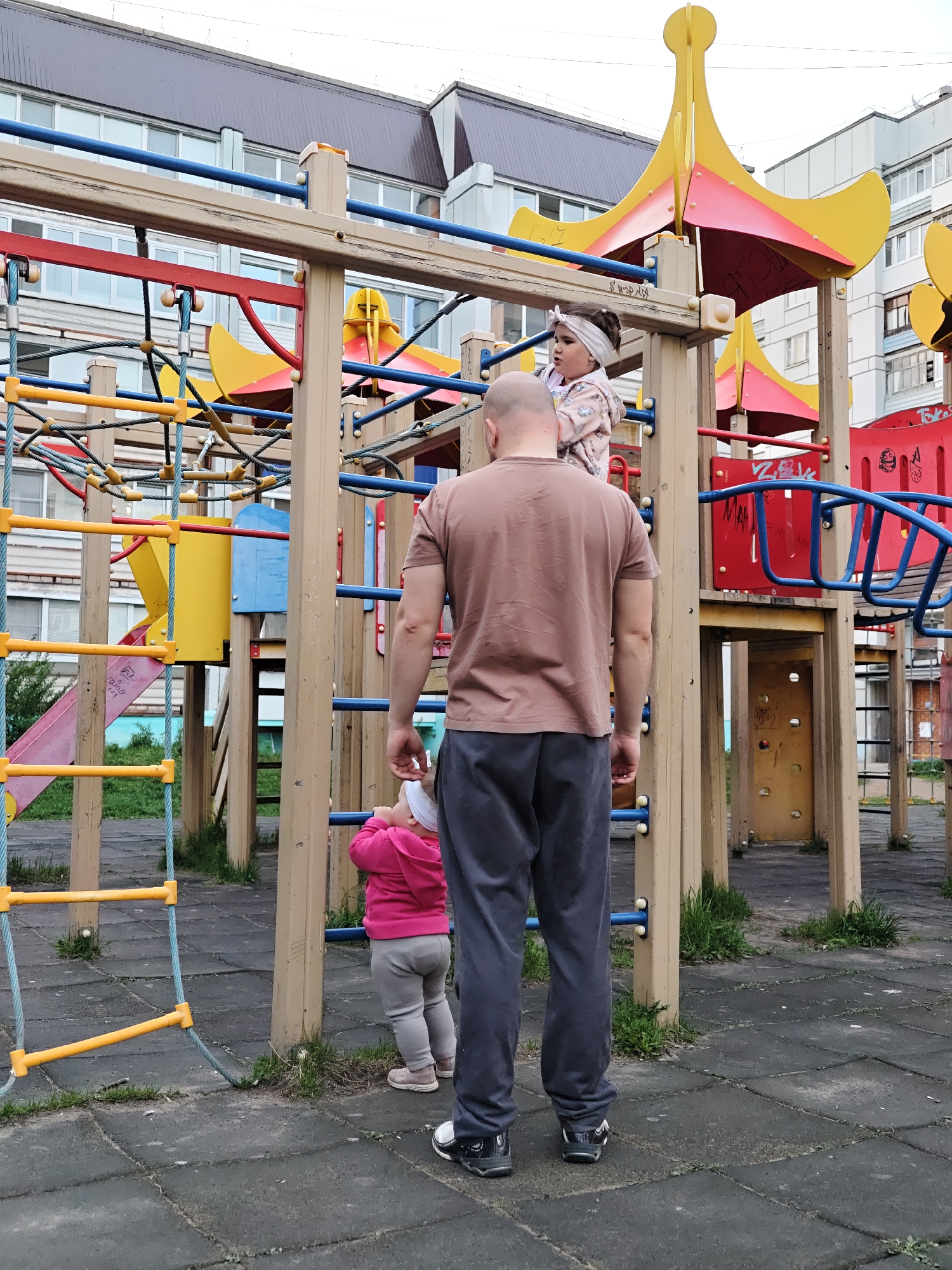 A playground in Chaikovsky, Russia, shows a bald man in a brown t-shirt and gray pants standing with his back to the camera, observing two young children. One child, wearing a patterned top and white headband, is standing on the climbing structure, while the other, in a pink jacket and white headband, is on the ground. The playground equipment is brightly colored with a mix of red, yellow, and blue, and includes slides, climbing bars, and rope ladders. The ground is paved with dark, textured rubber tiles. In the background, there are multi-story apartment buildings.