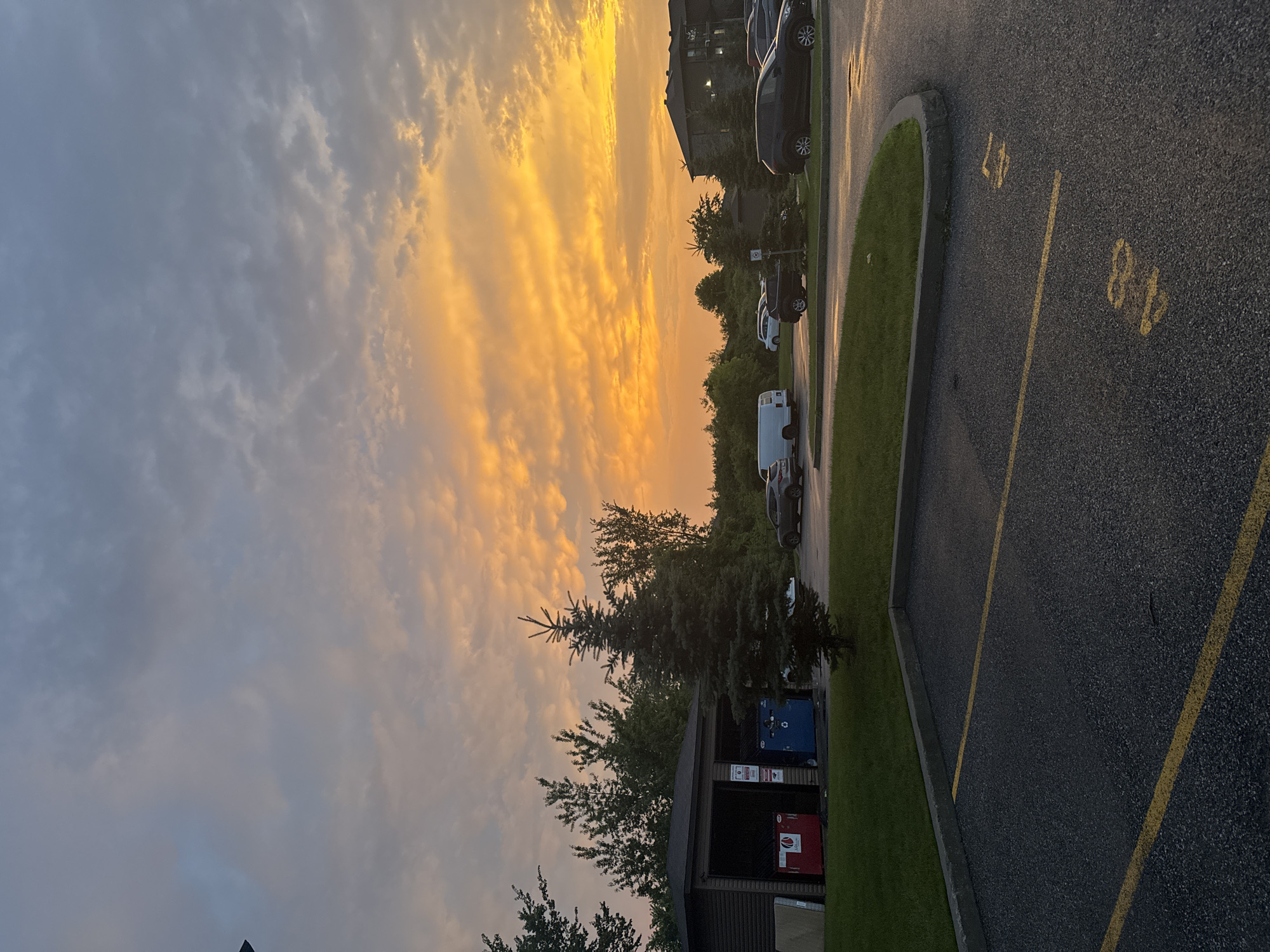 The image depicts an exterior scene at dusk in Gatineau, Canada. The sky is dominated by dramatic clouds with vibrant orange and yellow hues, indicative of a sunset. In the foreground, an asphalt parking lot is visible with yellow painted parking space markers, including the numbers "48" and "47". To the left, a small building with a dark roof and siding houses blue and red recycling bins. A young evergreen tree stands in front of this building. Behind the recycling station, several deciduous trees are sparsely leaved, suggesting summer. Further to the right, a multi-story building with a dark-pitched roof and stone-like siding is visible. Several cars, including a white van, a black SUV, and a dark sedan, are parked in the lot. The overall environment appears to be a residential or commercial complex. There are no people visible in the image.