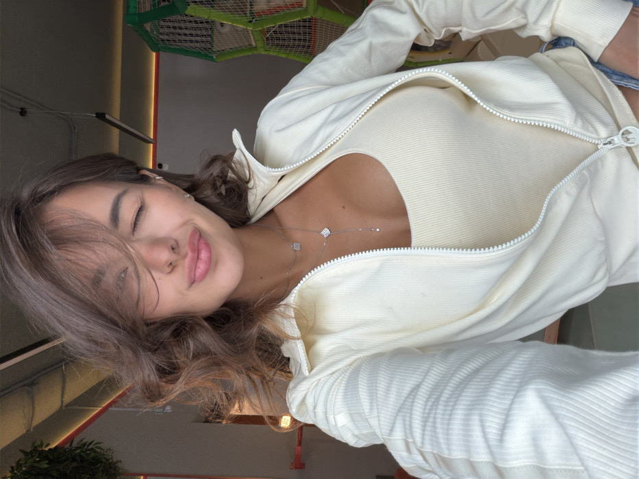 A young woman, with closed eyes and pursed lips, faces the camera. She has long brown hair and is wearing a white ribbed tank top and matching zip-up jacket, partially unzipped. A delicate silver necklace with clover-shaped pendants hangs around her neck. The background is slightly blurred and shows hints of a play area and interior setting.