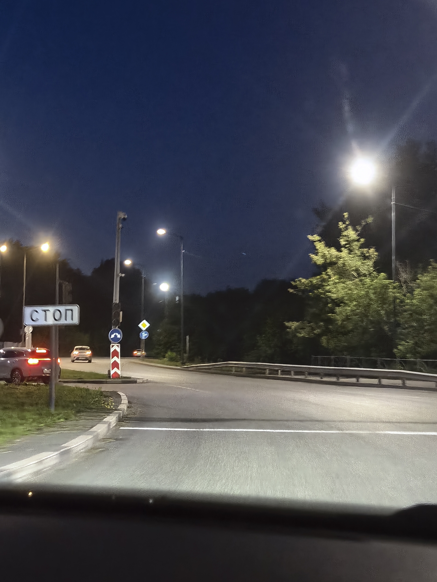 An eye-level, medium shot captures a nighttime scene at a road intersection. The sky is a deep, dark blue, illuminated by streetlights. The road surface is asphalt, with a white dividing line visible. On the left side of the road, a gray car is parked with its brake lights illuminated. A white sign with the Cyrillic text "СТОП" (STOP) is mounted on a pole beside the car. Further down the road, a light-colored car is in motion. A red and white chevron sign indicates a road barrier. A yellow diamond-shaped sign with a black border and a black symbol within is visible, along with a blue circular sign with a white arrow indicating a mandatory turn. Trees and foliage line the right side of the road, with the lights of street lamps casting a bright glow on them and the road. The scene appears to be on the outskirts of a city, possibly at a junction leading into or out of a wooded area. The perspective suggests the photo was taken from inside another vehicle, looking forward.
