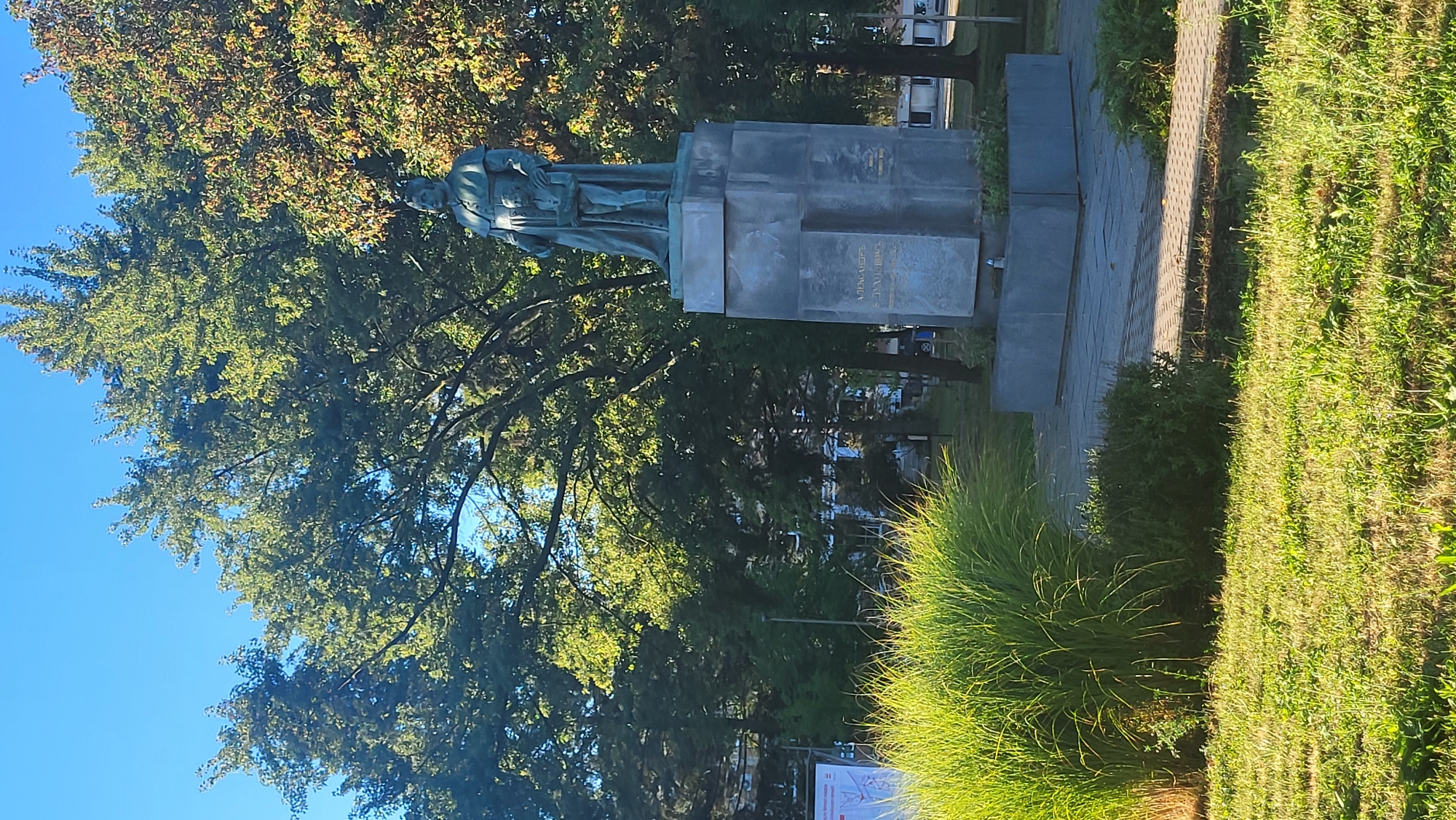 The image captures a bronze statue of a man in clerical attire, holding a child who is also holding a book. The statue stands on a tall, multi-tiered stone pedestal. The inscription on the pedestal is partially visible and appears to be in Cyrillic script. The statue is located in an urban park setting with lush green grass in the foreground and a variety of trees in the background, some with autumnal foliage. A paved walkway leads towards the statue. In the middle ground, there is a large, bushy ornamental grass. In the distance, buildings and parked cars are visible, suggesting a populated area. The sky is clear and blue.
This is the statue of Orthodox Bishop of Mukačevo and Prešov, Dositej (or Dosifei) Poopovich, located in the park on Štefánikova Street in Prešov, Slovakia.