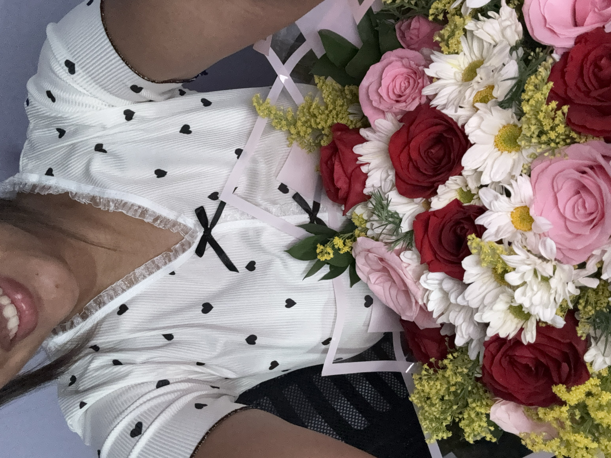 A woman with dark hair is holding a bouquet of red and pink roses, as well as white daisies, up to her face. She is wearing a white pajama top with black heart designs and lace trim. Her lips are visible in the lower left corner of the frame. The bouquet is wrapped in a white geometric-patterned paper. The background is a light blue color. The location is not specified in the image.