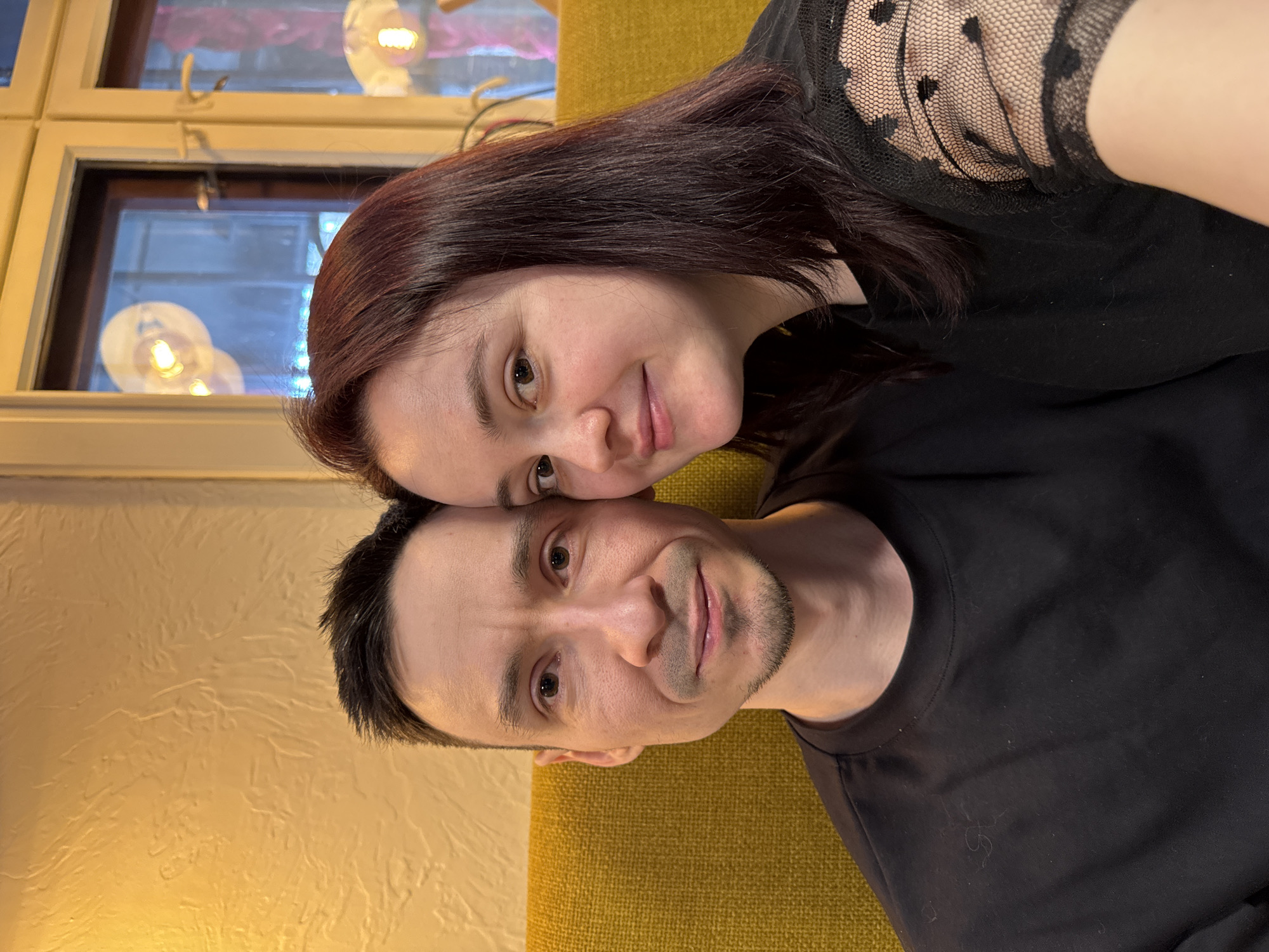 A couple is posing for a photo. The woman has dark red hair and is wearing a black lace top. She is leaning her head on the man's head. The man has short black hair and is wearing a black t-shirt. They are sitting on a mustard yellow couch. A window with light fixtures reflecting in it is visible in the background. The location is Izhevsk, Russia.