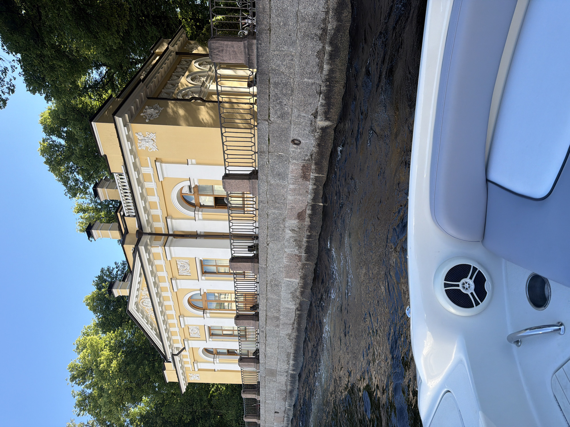 The image shows a boat in a canal next to a yellow building with a neoclassical facade. The building has decorative carvings, arched windows, and a black wrought-iron fence along the canal. Lush green trees are visible behind the building. The water in the canal is dark and choppy. The boat's interior, featuring white and light blue upholstery and a speaker, is in the foreground on the right side of the image. The scene appears to be set during the daytime, with clear blue skies indicating good weather. The perspective suggests the photo was taken from the boat, looking towards the shore. The overall mood is serene, capturing a leisurely moment on the water. There is no visible text in the image.
