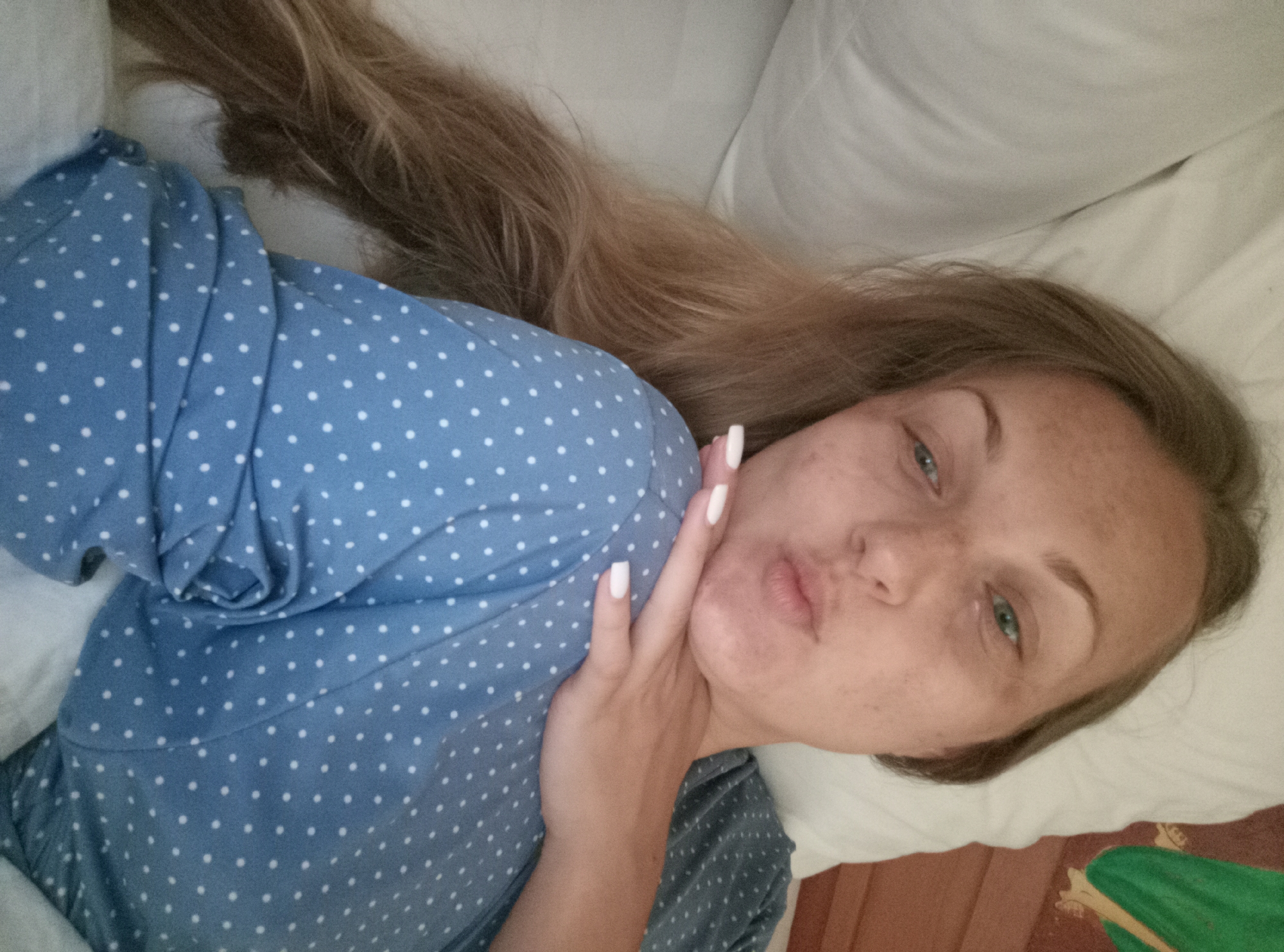 A woman with long blonde hair and fair skin lies on a bed with white pillows and a wooden headboard. She is wearing a blue polka-dot pajama top and has white-painted fingernails. She is puckering her lips and resting her chin on her hand. The lighting appears to be natural. The scene is indoors and appears to be a bedroom. There is no indication of the location being in Denpasar, Indonesia.