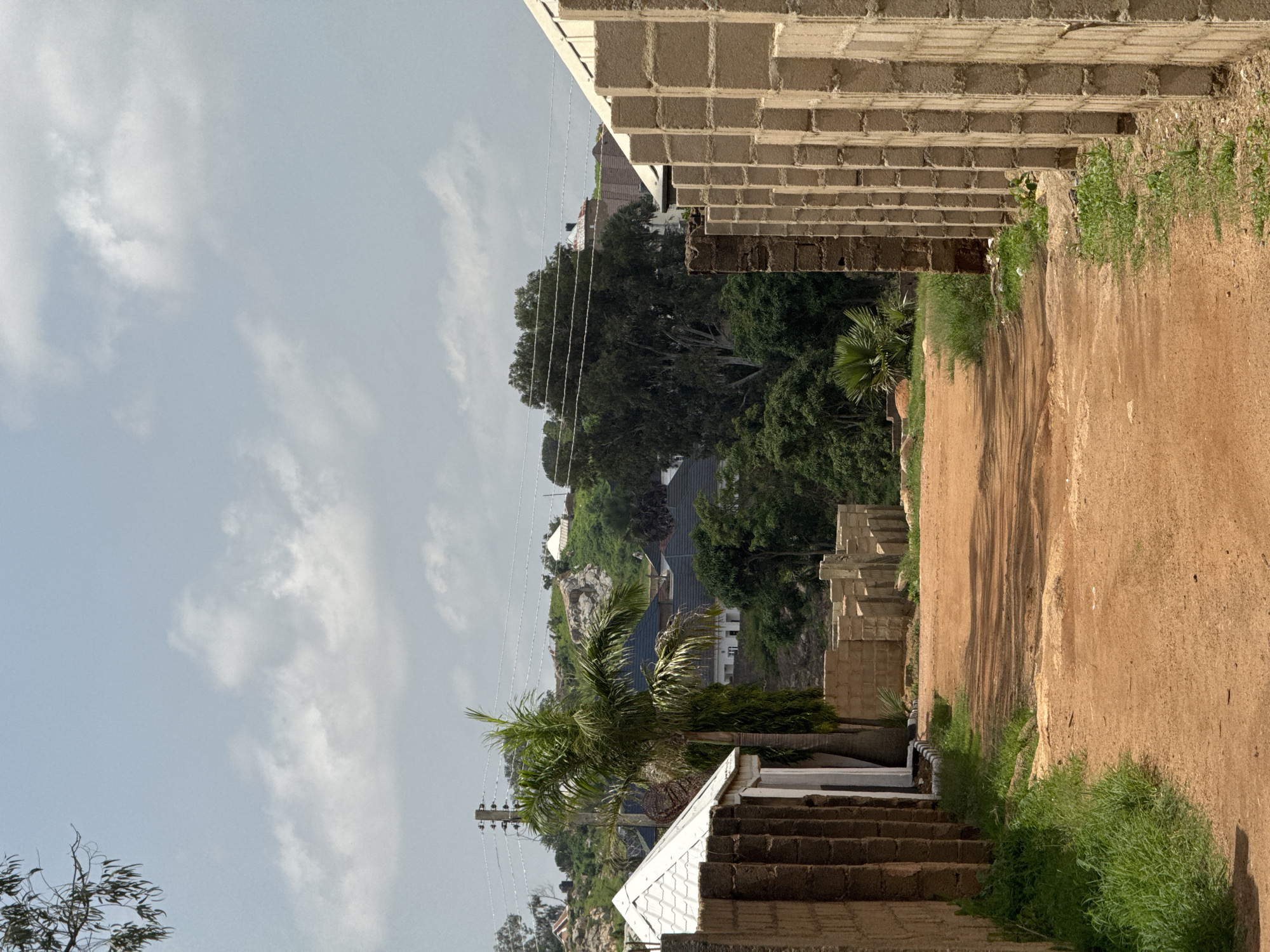 This is a vertical, outdoor, eye-level, medium shot of a hilly, treed landscape in Gura Top, Nigeria, under a bright, cloudy sky. The foreground is dominated by a rough, reddish-brown dirt path running diagonally from the bottom right to the upper left of the frame. To the left of the path, a low wall constructed of light-colored cinder blocks lines the edge of a building in the process of construction. The wall extends into the upper left of the image. Green grass and weeds grow along the base of the wall and the edge of the path.
In the mid-ground, a dense forest of lush green trees covers the rolling hills. Among the trees, glimpses of buildings with dark roofs and white walls are visible. A tall palm tree with its fronds spread wide stands prominently in the middle left of the frame. Electrical wires stretch across the scene, supported by a utility pole.
Further back, the hills rise, showcasing more vegetation and scattered houses. The sky is a light blue with scattered, white, fluffy clouds. The lighting suggests it is daytime, likely late morning or early afternoon, given the brightness and some of the shadows cast. The overall scene depicts a developing area with ongoing construction alongside natural landscapes. No people are visible, and no text is present in the image.