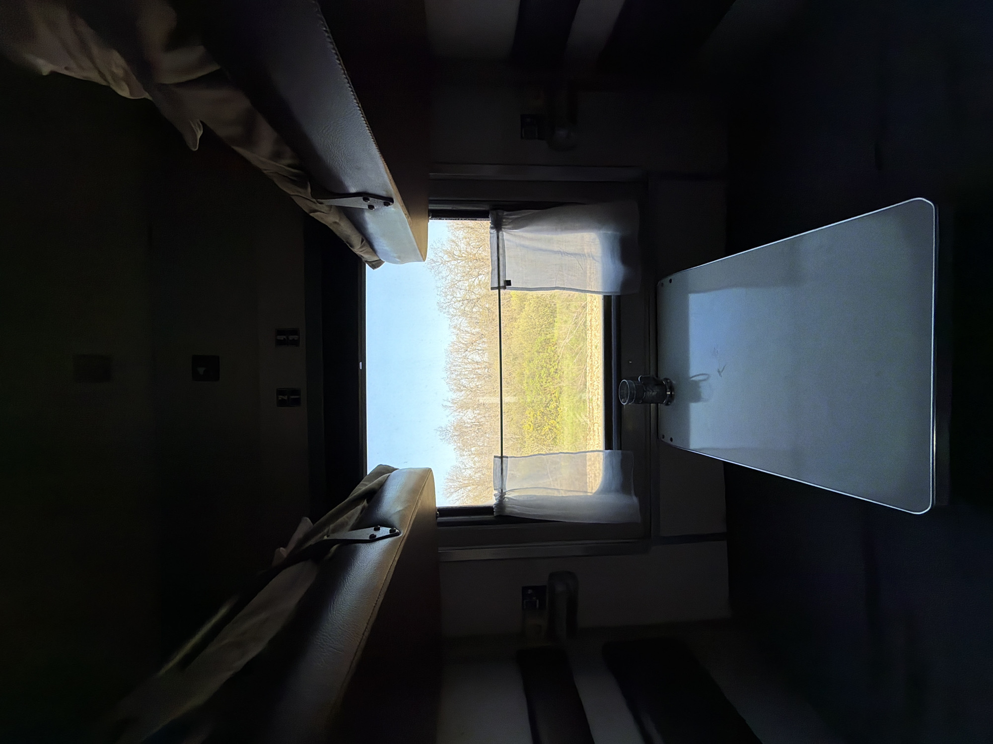 The image shows the interior of a train compartment. There are two bunk beds visible, one above the other. A window with sheer curtains reveals a view of trees and greenery, suggesting a rural landscape. A small, foldable table is attached to the wall on the right side of the window. The lighting is dim, primarily coming from the window. There is no evidence that this is in Lebyazh'e, Russia.
