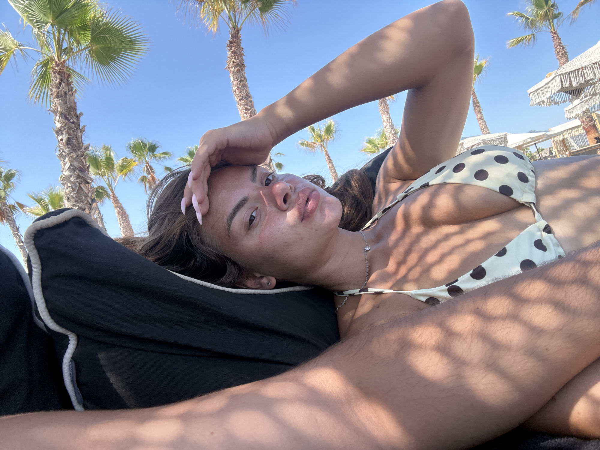 A young woman is lying on a lounge chair outdoors, with palm trees and clear blue sky in the background. She is wearing a polka dot bikini. Her hand is resting on her forehead, and she is looking towards the camera with a relaxed expression. The scene appears to be a beach or resort, given the palm trees and lounge chairs. The lighting suggests it is daytime, likely sunny and warm. There are some shadows cast on her and the chair from overhead foliage or structures, indicating a bright, sunny day. The woman has long, dark hair and fair skin.