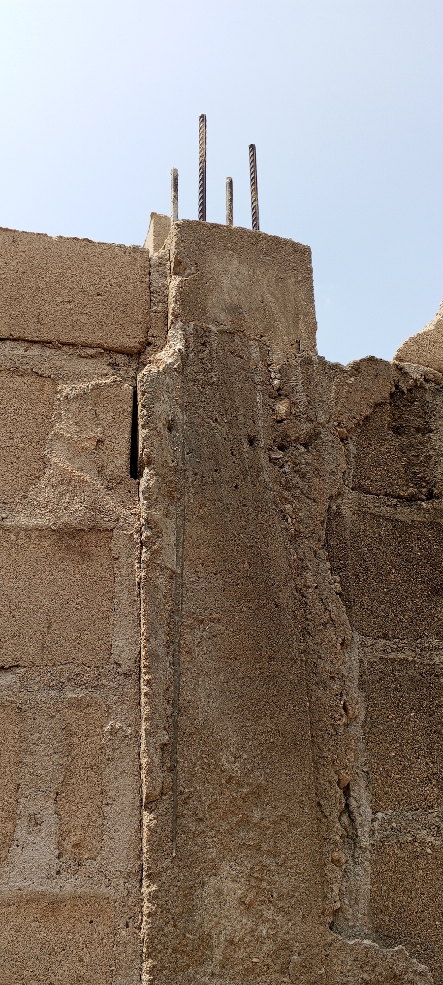 The image shows the top of a concrete structure under construction, with four rebar rods protruding upwards against a clear sky. The concrete appears rough and textured, with some visible aggregate. The structure is composed of large, roughly finished concrete blocks. A narrow, dark vertical gap is visible on the left side of the structure. The scene is likely located in Jalingo, Nigeria, given the architectural style and apparent construction activity common in the region. There are no people or animals visible in the image. The lighting suggests it is daytime, with the sun casting shadows that are not prominently visible due to the angle. The overall impression is of an unfinished building element.