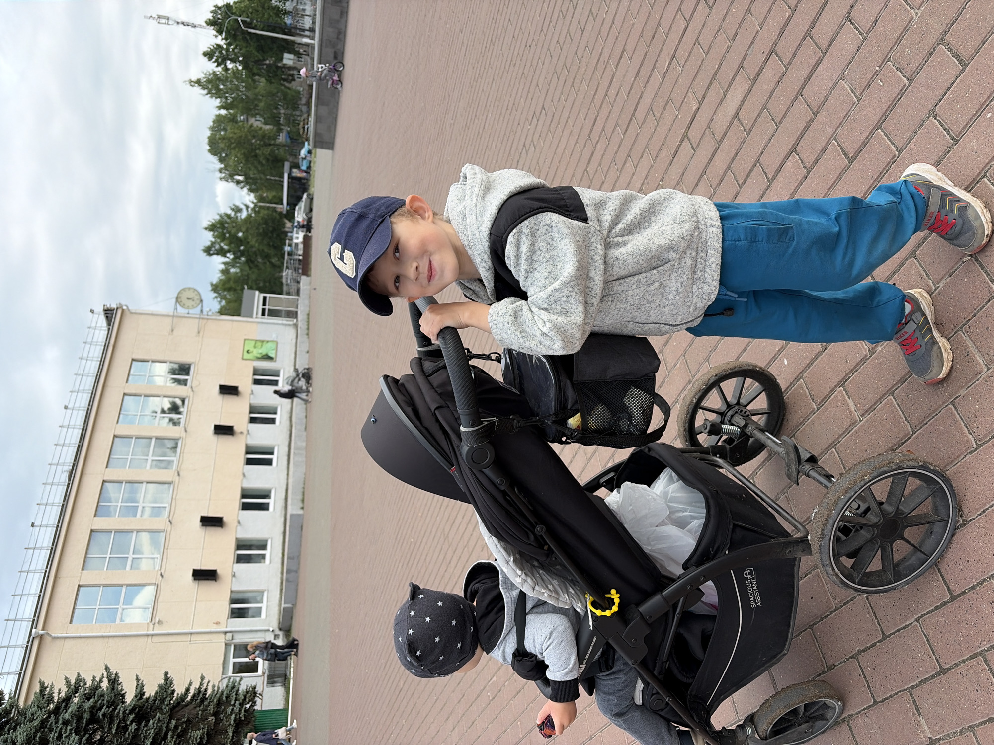 The image captures an outdoor scene in Chaikovsky, Russia, featuring two children on a paved walkway during what appears to be an overcast daytime.
The primary subjects are an older boy and a younger child in a stroller. The older boy, positioned on the right, is looking at the camera with a smile. He wears a navy blue baseball cap with a light-colored 'C' or 'G' logo, a grey and black hooded jacket, blue trousers, and grey sneakers with red laces, featuring "BIKER" written on the strap. He is leaning on the handle of a black stroller.
The younger child is seated in the stroller, visible from the back. They wear a black hat adorned with white stars and a grey hooded outfit. A yellow teether or toy is clipped to the stroller's handle near them. The robust black stroller has large, somewhat muddy wheels, and a mesh pocket on the side contains a yellow ball or toy. Text "SPACIOUS ASSISTANT" and "ALL SEASONS READY" are visible on the stroller's lower basket. A white plastic bag is also seen inside the storage area.
The setting is an urban or suburban area with a reddish-brown brick-paved ground. In the background, to the left, stands a light-colored multi-story building with numerous windows and a large round clock on its facade. Below the clock is a green square sign. Further back, more trees and taller buildings are visible, along with a communication tower. The sky is largely overcast, suggesting a mild, cloudy day. The older boy's expression conveys a happy or playful mood.