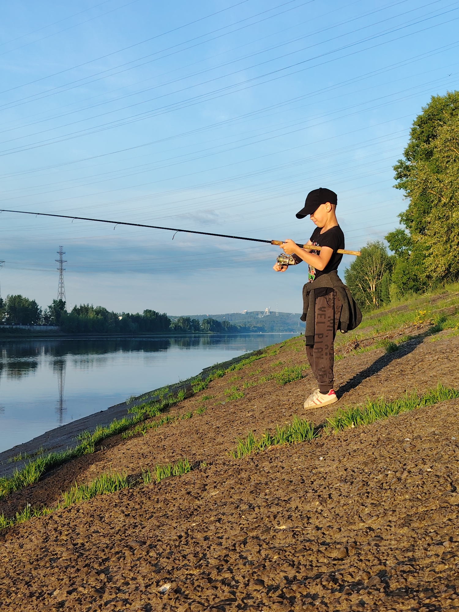 A young boy, wearing a black t-shirt, black baseball cap, and dark pants with a grey jacket tied around his waist, stands on a dirt embankment with sparse green grass. He is holding a fishing rod, with the line extending over a calm body of water. The boy is positioned to the right of the frame, facing left. The water reflects the clear blue sky, which is crisscrossed by power lines. In the distance, across the water, is a treelined shore and a city skyline with industrial structures, including a tall transmission tower. The sun appears to be low in the sky, casting long shadows and illuminating the scene with warm light. The location is the Kama River embankment in Chaikovsky, Russia.