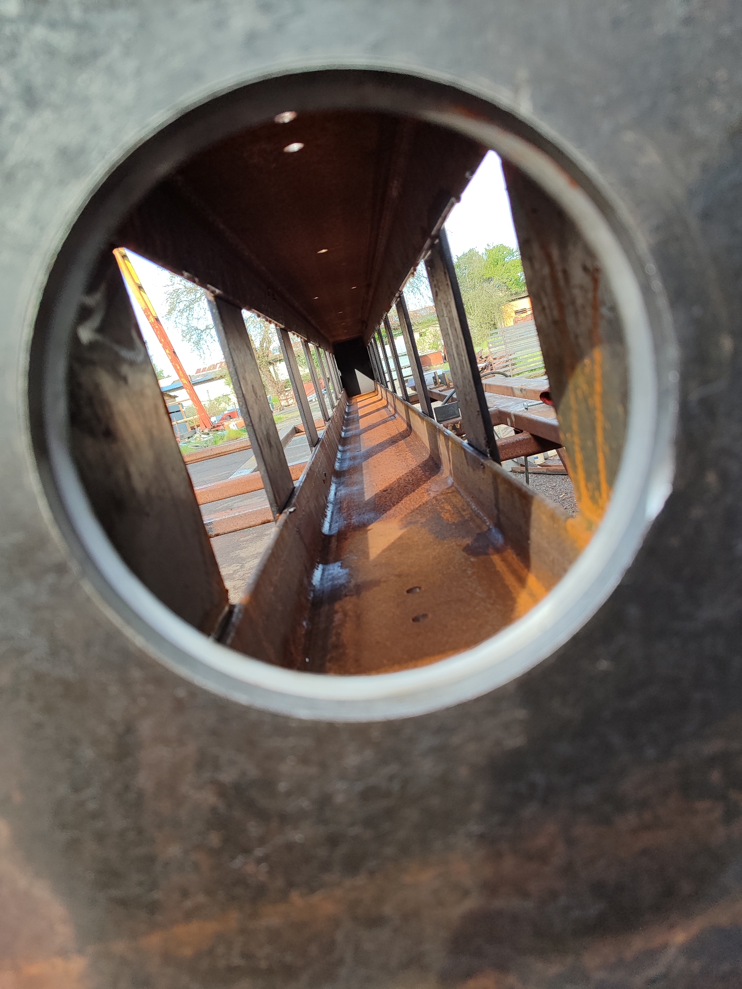 The image presents a perspective shot looking through a circular opening, possibly a pipe or a large bolt hole, into a long, open-top metal channel or trough. The circular foreground element is made of dark, textured metal with a lighter, smooth inner rim.
Inside the channel, the dominant subject is the rusty, reddish-brown metal structure. The channel extends far into the distance, creating a strong sense of depth. Its bottom surface appears wet or reflective, possibly from water or oil, catching the light and creating bright streaks. Several vertical metal beams or supports are visible within the channel, particularly along its left side, suggesting a robust construction. Small, bright points of light are scattered across the upper inner surface of the channel, possibly reflections or imperfections.
Beyond the end of the channel, an outdoor scene is visible under clear daylight. Green foliage from trees can be seen, along with parts of buildings, one of which appears to have a reddish roof. On the left, a large red structure, possibly a crane or construction equipment, is partially visible. The overall setting suggests an industrial yard or construction site in Burgas, Bulgaria.
No people are present, and no specific activity is taking place; the image captures a static view of industrial infrastructure. The bright lighting indicates it is daytime, possibly morning or late afternoon, given the shadows and light reflections within the channel. No visible text is present in the image.