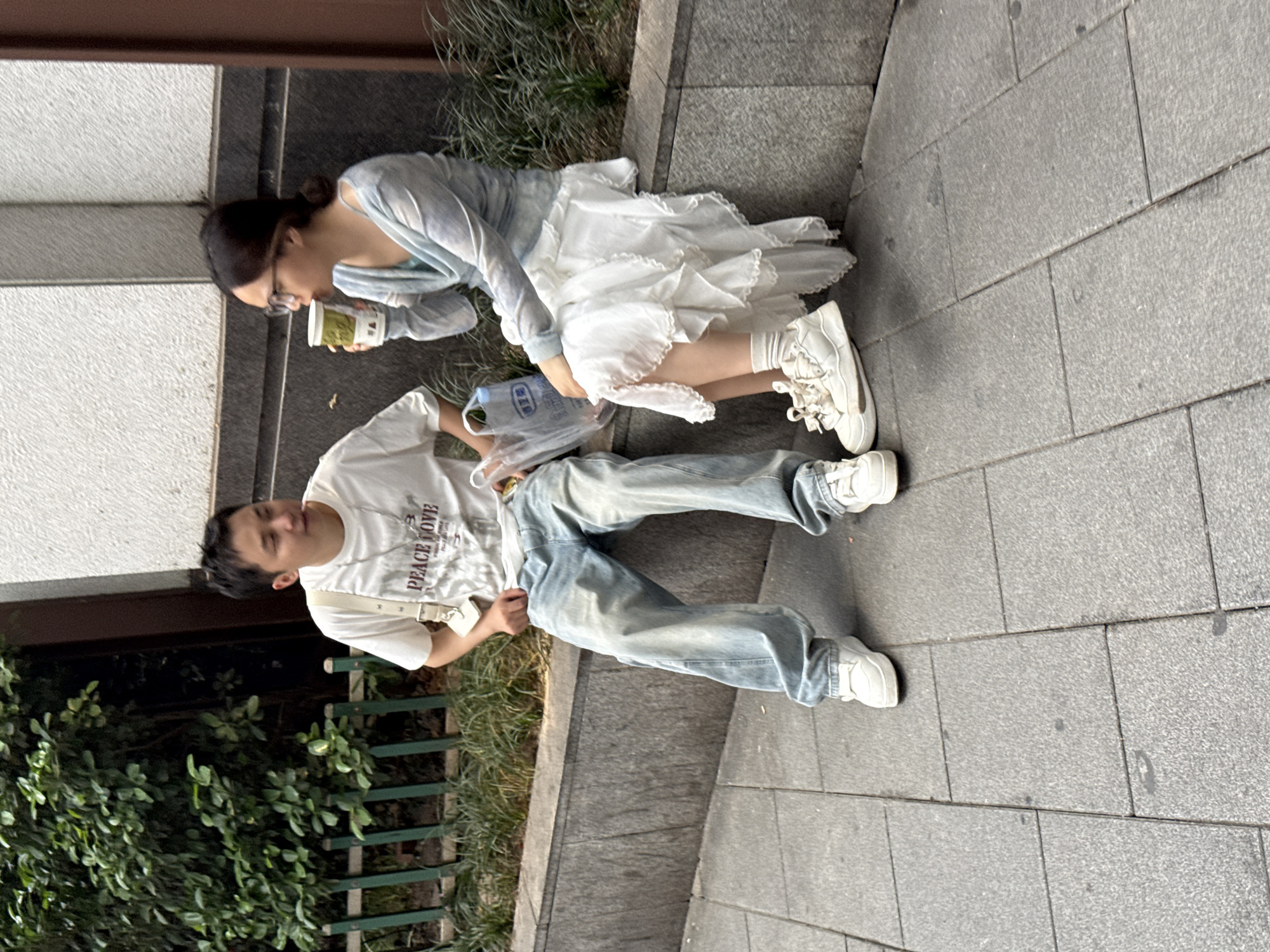 Two people are sitting on a sidewalk in Yuzhong District, China. A woman in a light blue top and white dress sits near a building, holding a beverage. A man in a white t-shirt and blue jeans sits adjacent to her on the sidewalk. A plastic bag rests between them. The sidewalk is made of gray bricks, and green plants grow nearby.