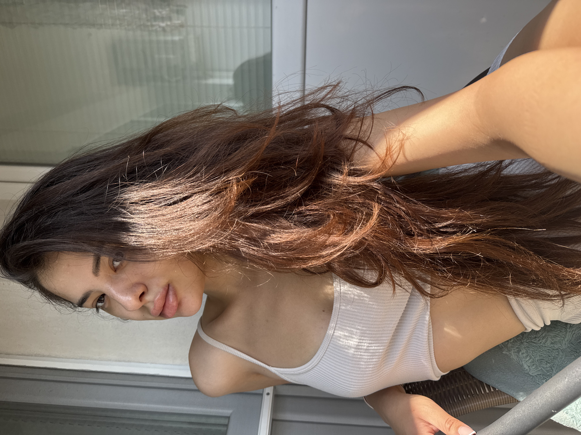 An eye-level medium close-up photograph captures a young adult woman seated outdoors in the city of Plovdiv, Bulgaria, likely on a balcony or patio. She has long, wavy, dark brown hair with reddish undertones, which falls over her right shoulder and chest, partially obscuring her right arm. Her head is turned towards the viewer, and her dark eyes are looking directly at the camera. She has full, glossy lips, a prominent nose, and an olive skin tone. Her complexion shows natural texture, with some minor blemishes visible.
She is wearing a light-colored, possibly off-white or beige, ribbed spaghetti-strap crop top that reveals a sliver of her midriff. Below the crop top, a hint of matching light-colored fabric, possibly shorts or a skirt, is visible. Her left arm is bent at the elbow, with her hand resting on the armrest of a wicker chair. Her fingernails have a French-tipped manicure. Her right arm is bent with the forearm resting on her lap, largely covered by her hair.
Strong, directional sunlight from the upper right illuminates the right side of her face, shoulder, and hair, creating bright highlights and distinct shadows across her body and the background.
Behind her is a plain, light-colored wall (off-white or pale gray). To her left (viewer's right) is a window or glass door with horizontal blinds, reflecting ambient light. A narrow vertical window frame or door jamb is visible on the far left edge of the frame. In the lower left foreground, a gray metal railing or part of the chair structure is visible. A light blue or teal textured fabric is draped over the chair armrest beneath her left hand.